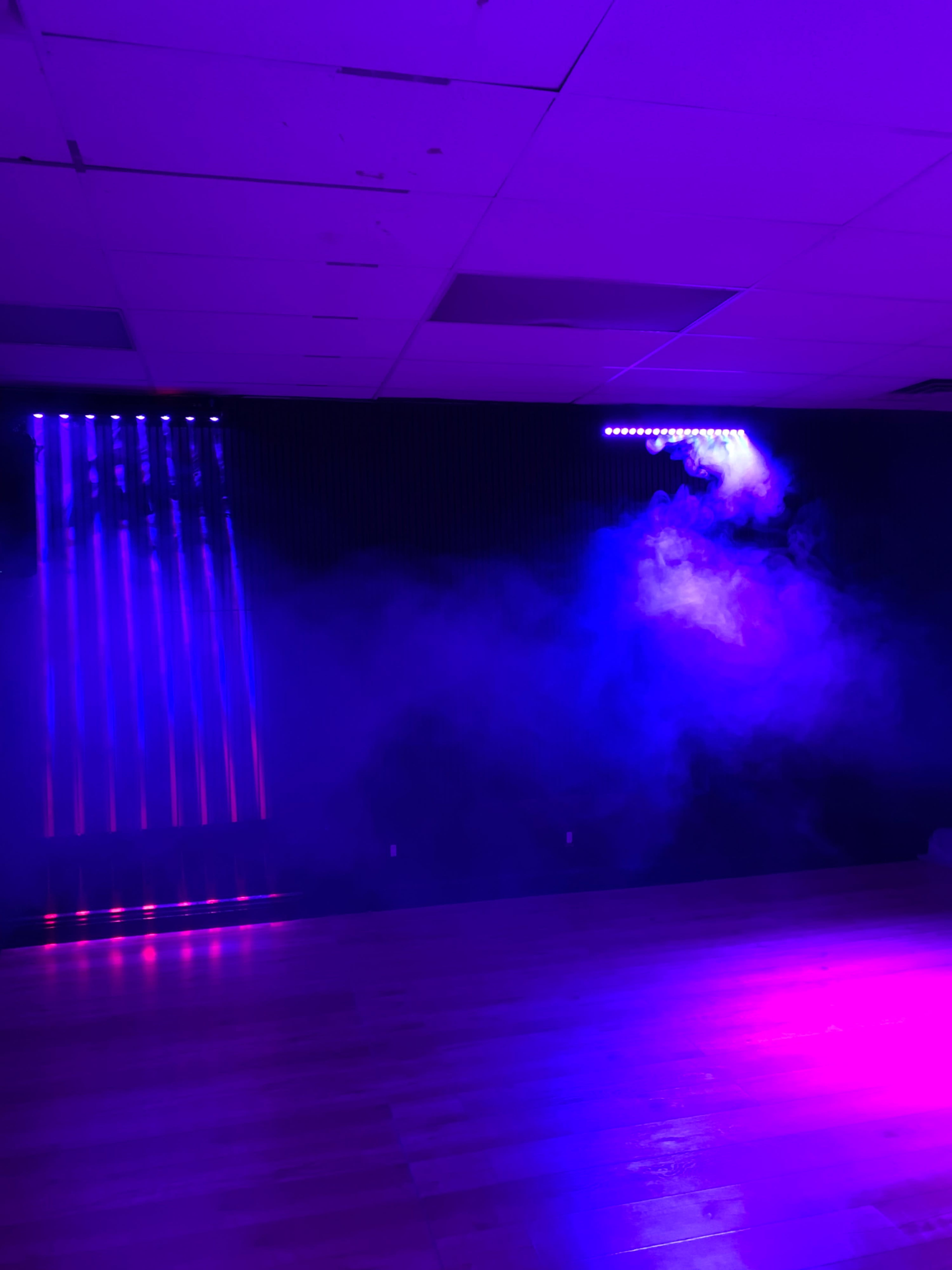 A dimly lit room features colorful lights casting beams through a haze of fog.