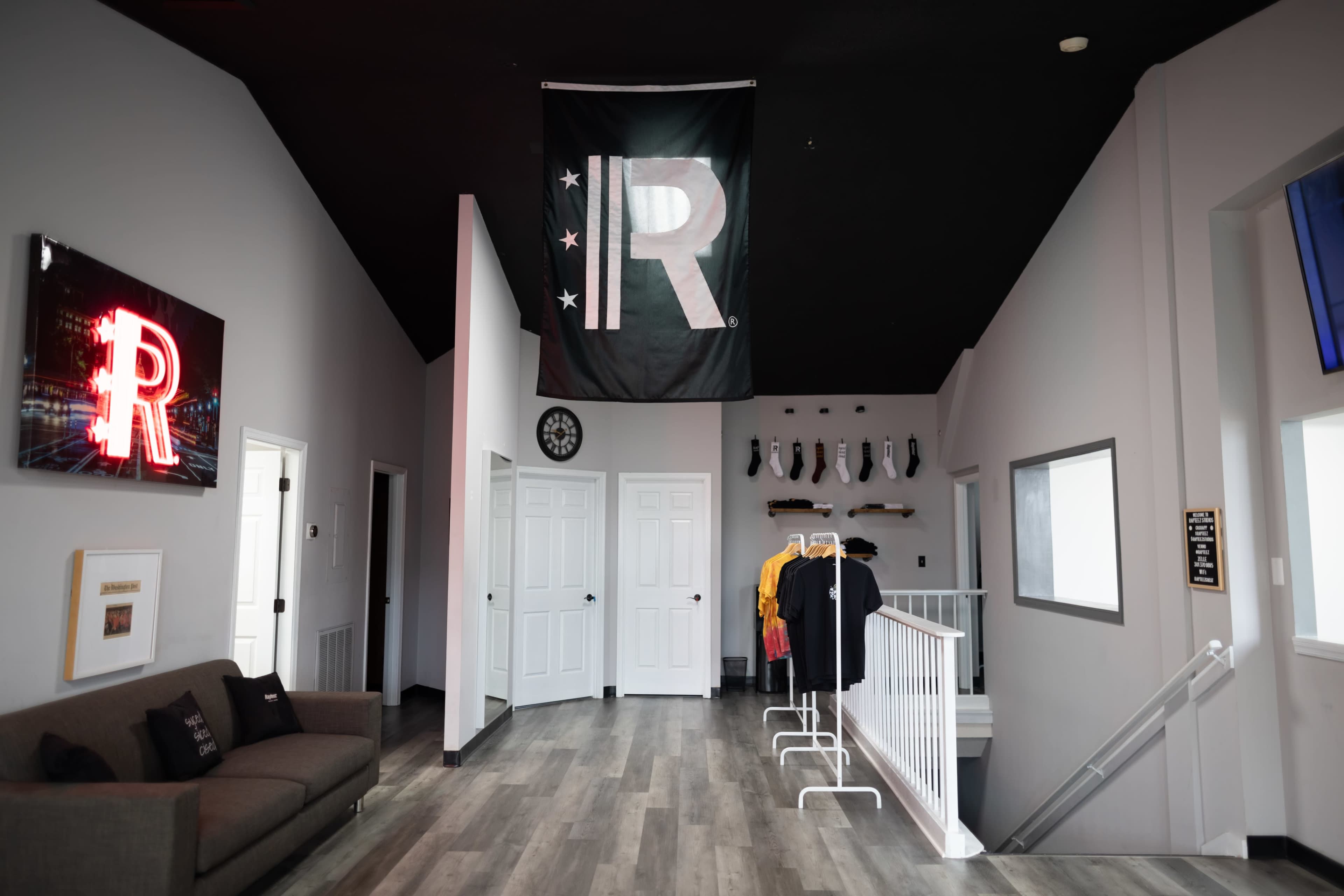 The image shows a modern interior space with a large black flag featuring the letter "R" hanging from the ceiling, a gray sofa on the left, and clothing displayed on a rack in the corner.