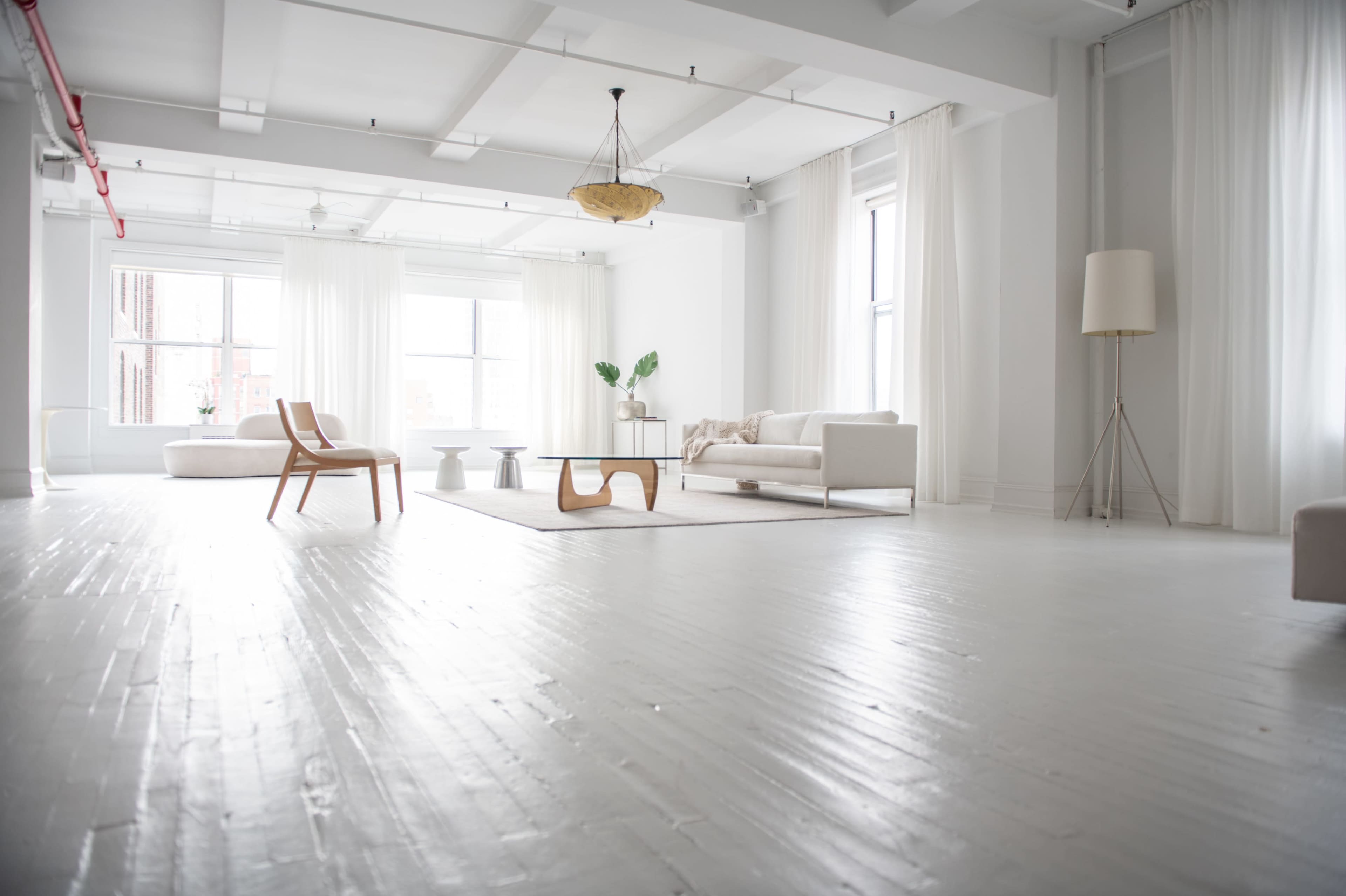 A spacious, white-walled room features minimalist furniture, large windows with sheer curtains, and a light wooden floor.
