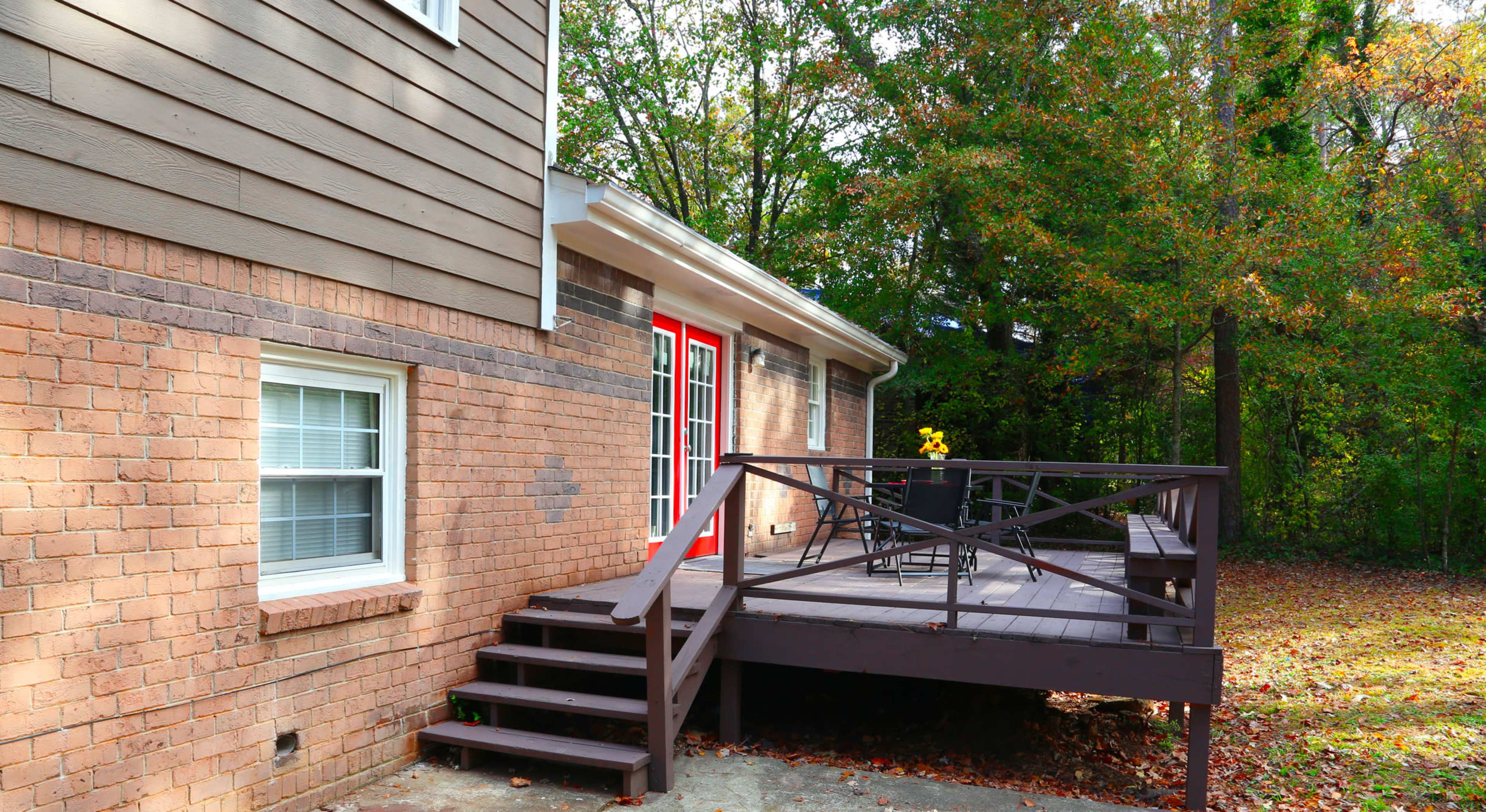 A brick house with a wooden deck and steps leading to an outdoor seating area surrounded by trees.
