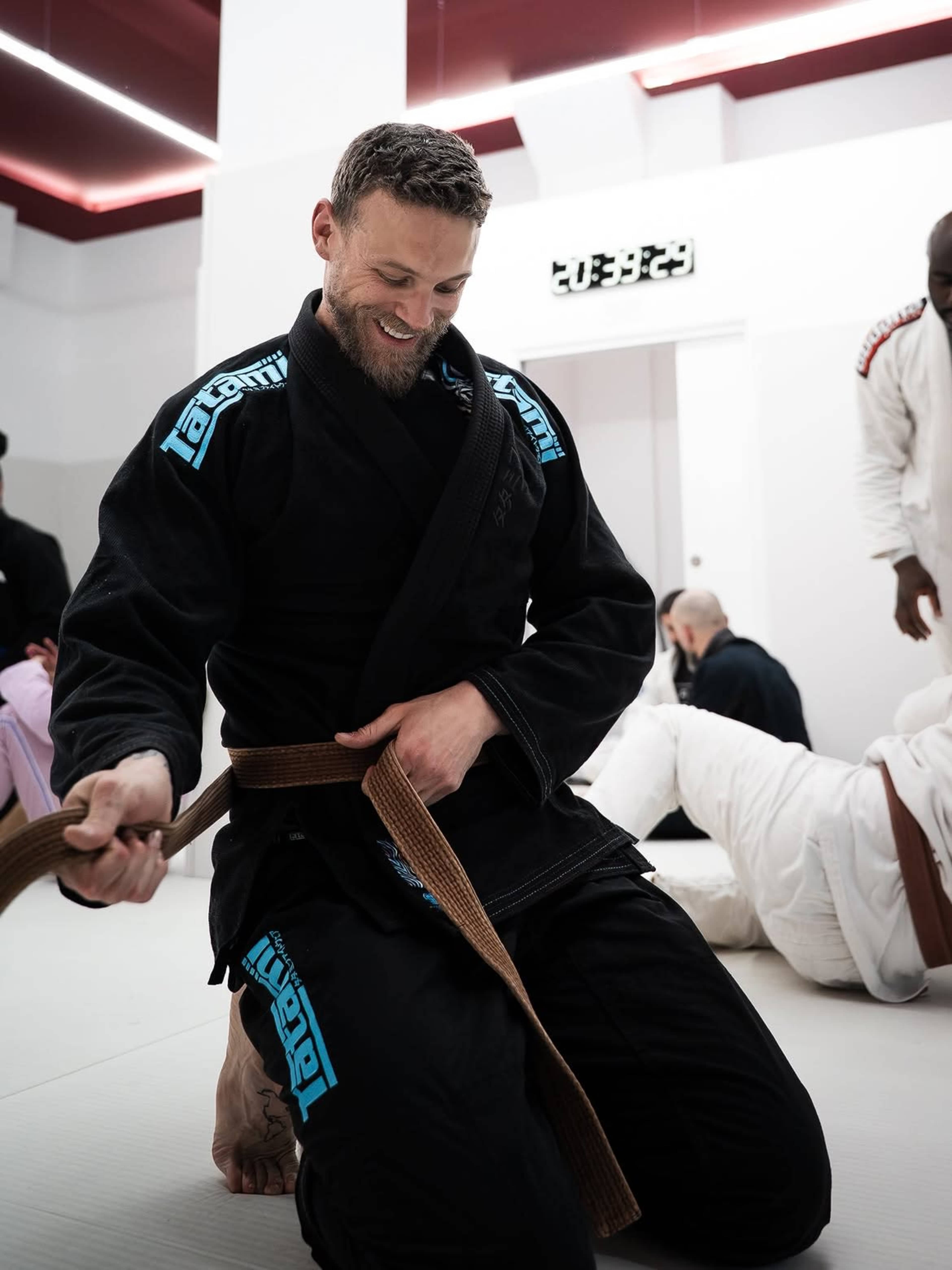 A man in a black Brazilian jiu-jitsu gi is kneeling with a brown belt in a training gym, surrounded by other practitioners engaged in various activities.
