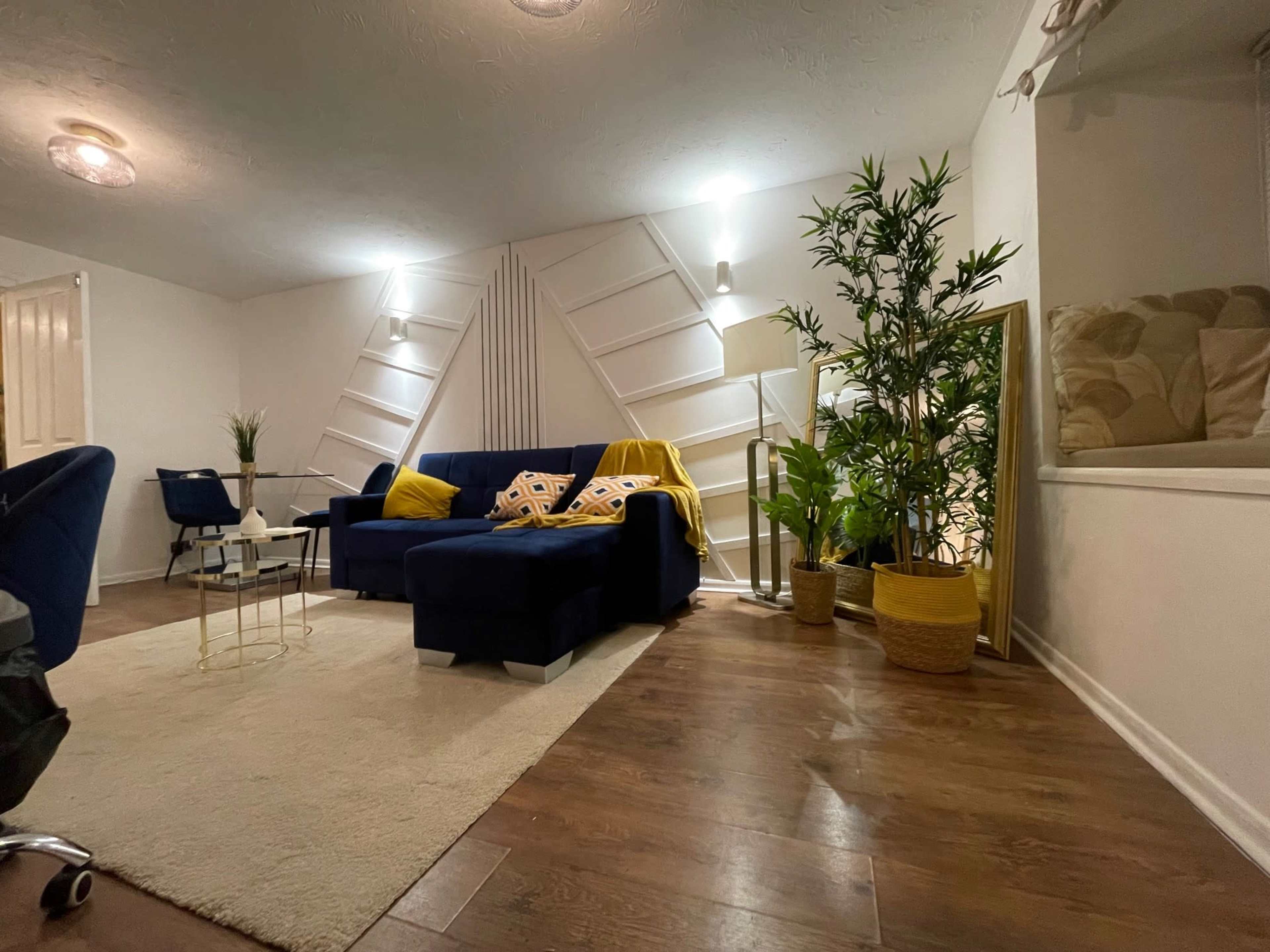 A modern living room features a blue sofa with decorative cushions, a coffee table, indoor plants, and a large mirror against a white wall.