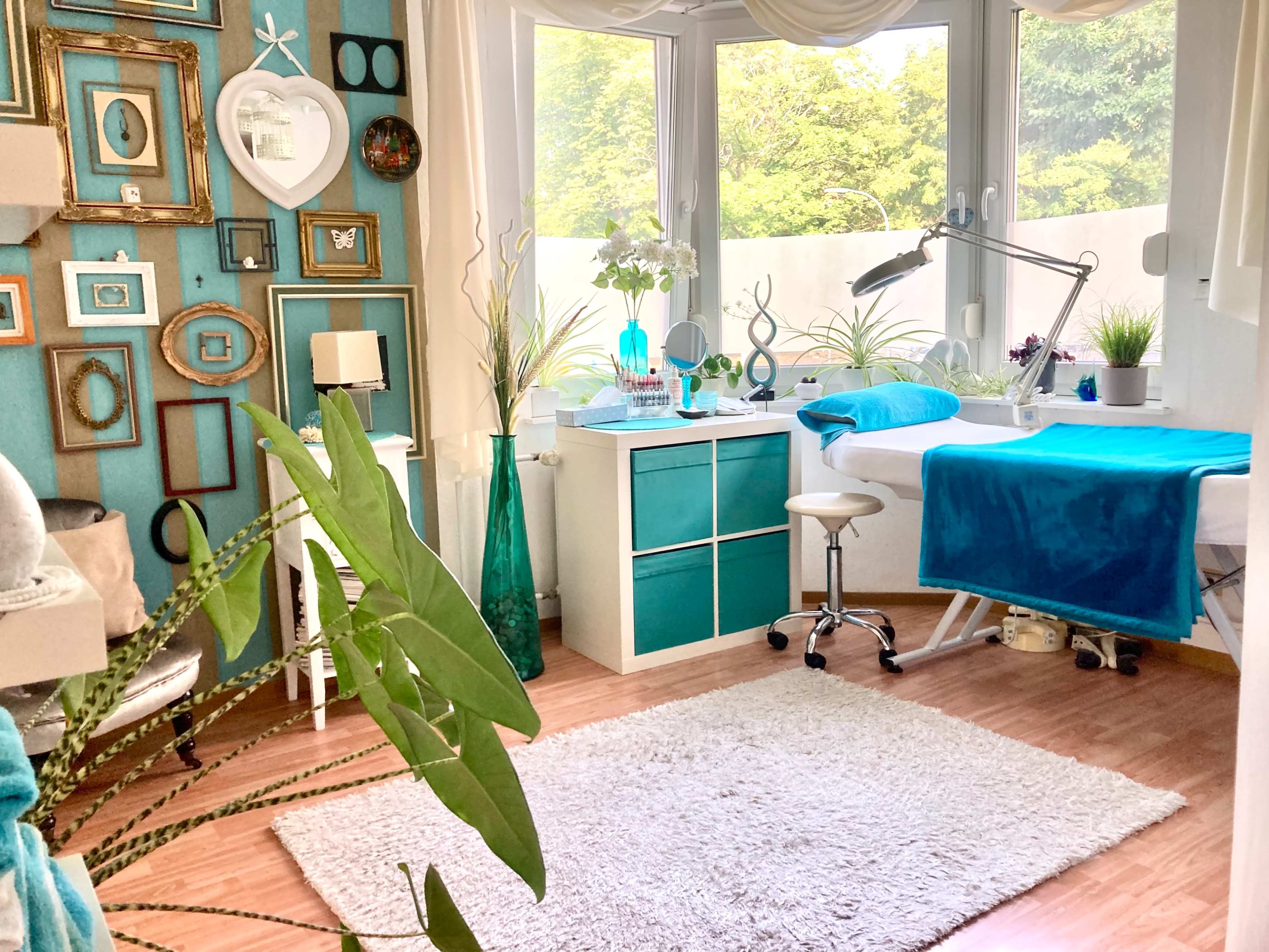 The image shows a bright, organized room with teal accents, a treatment table, decorative plants, and a desk area surrounded by framed artwork.