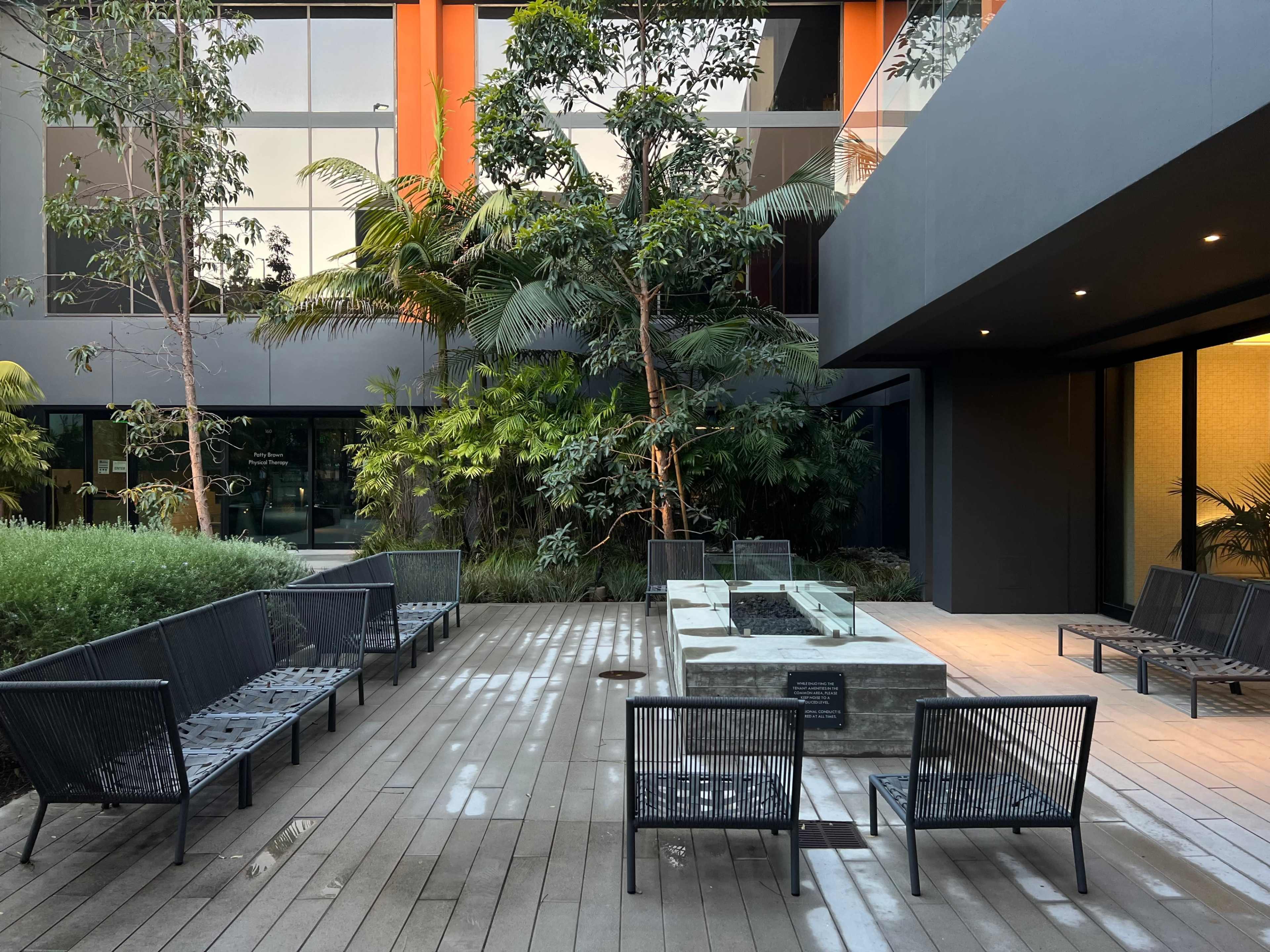The image shows a modern outdoor seating area with black metal chairs surrounding a rectangular concrete fire pit, set amidst lush greenery and contemporary architectural elements.