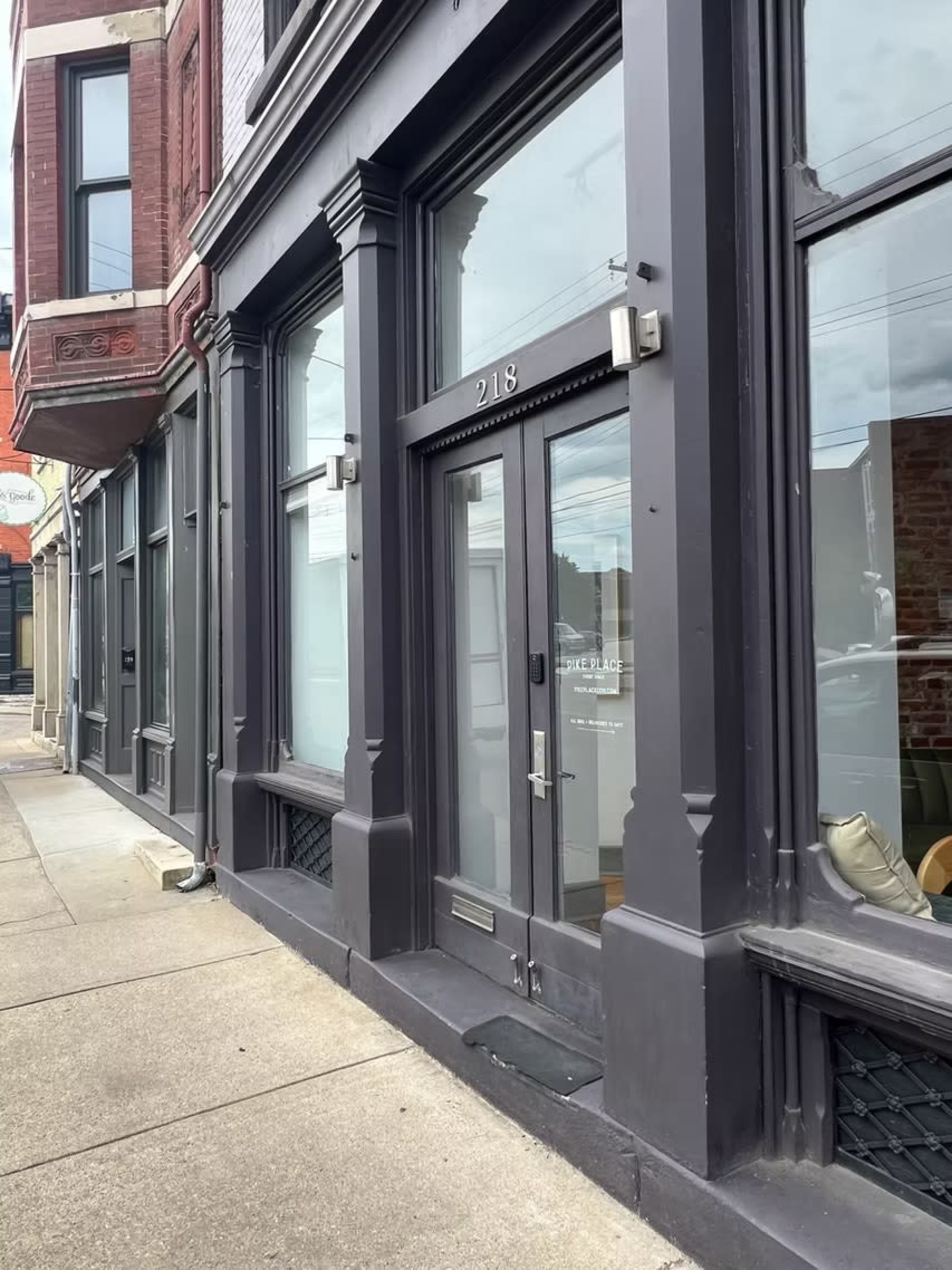 The image shows a gray storefront with large windows and a glass door, located on a sidewalk next to adjacent buildings.