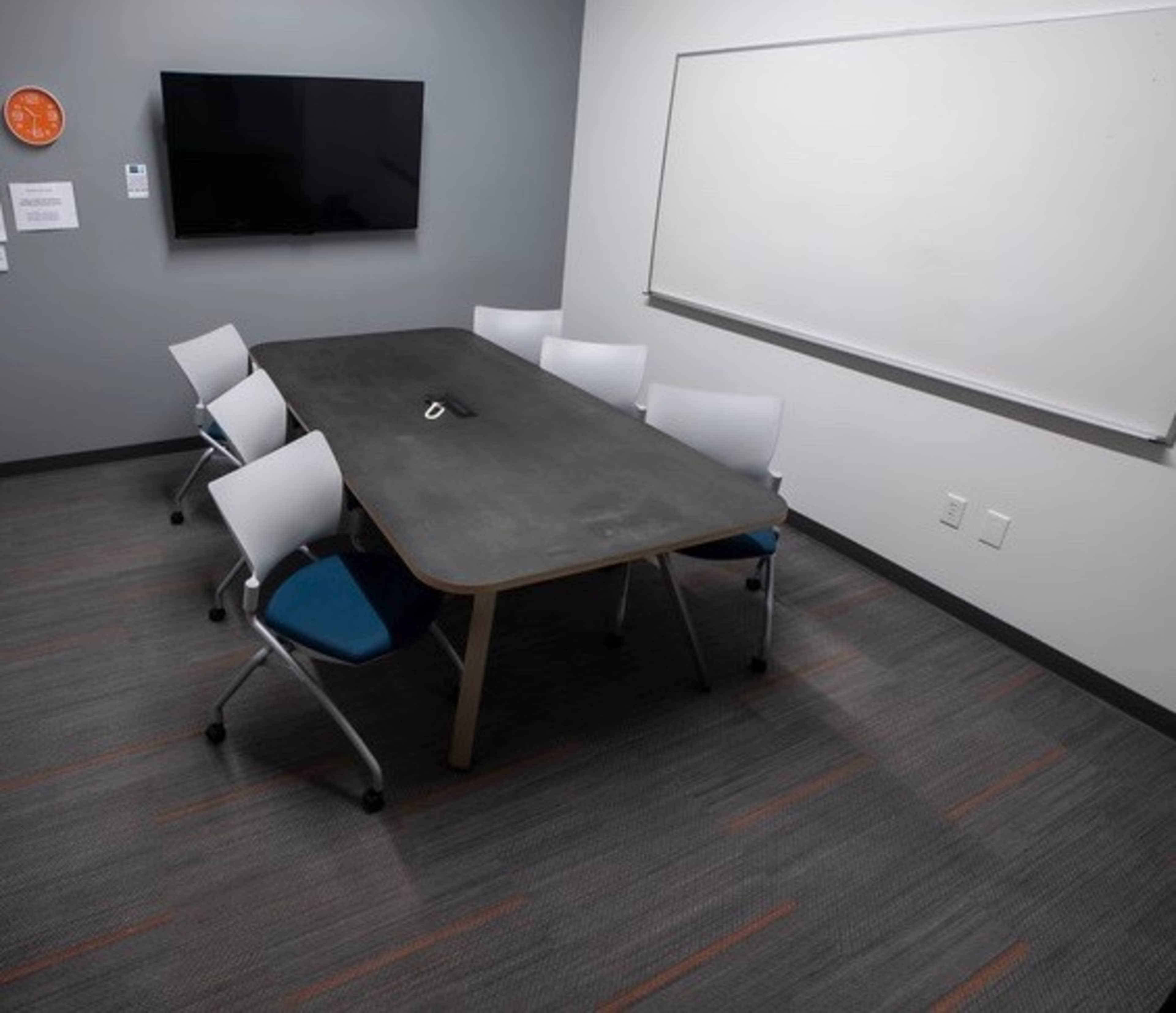 The image shows a conference room with a large table surrounded by six chairs, a wall-mounted television, and a whiteboard.