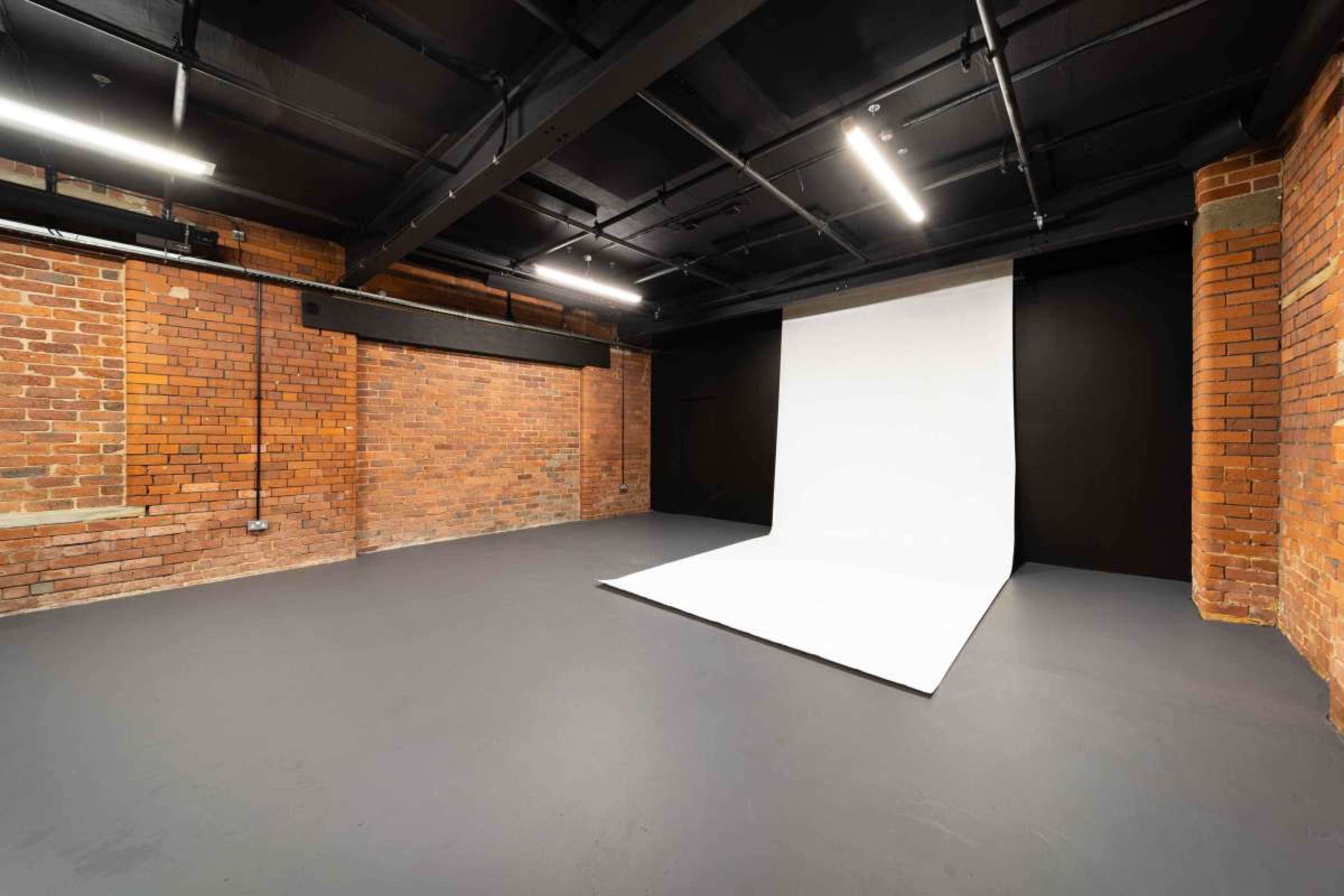 The image shows an empty studio space with exposed brick walls, a dark ceiling, and a white backdrop on the floor.