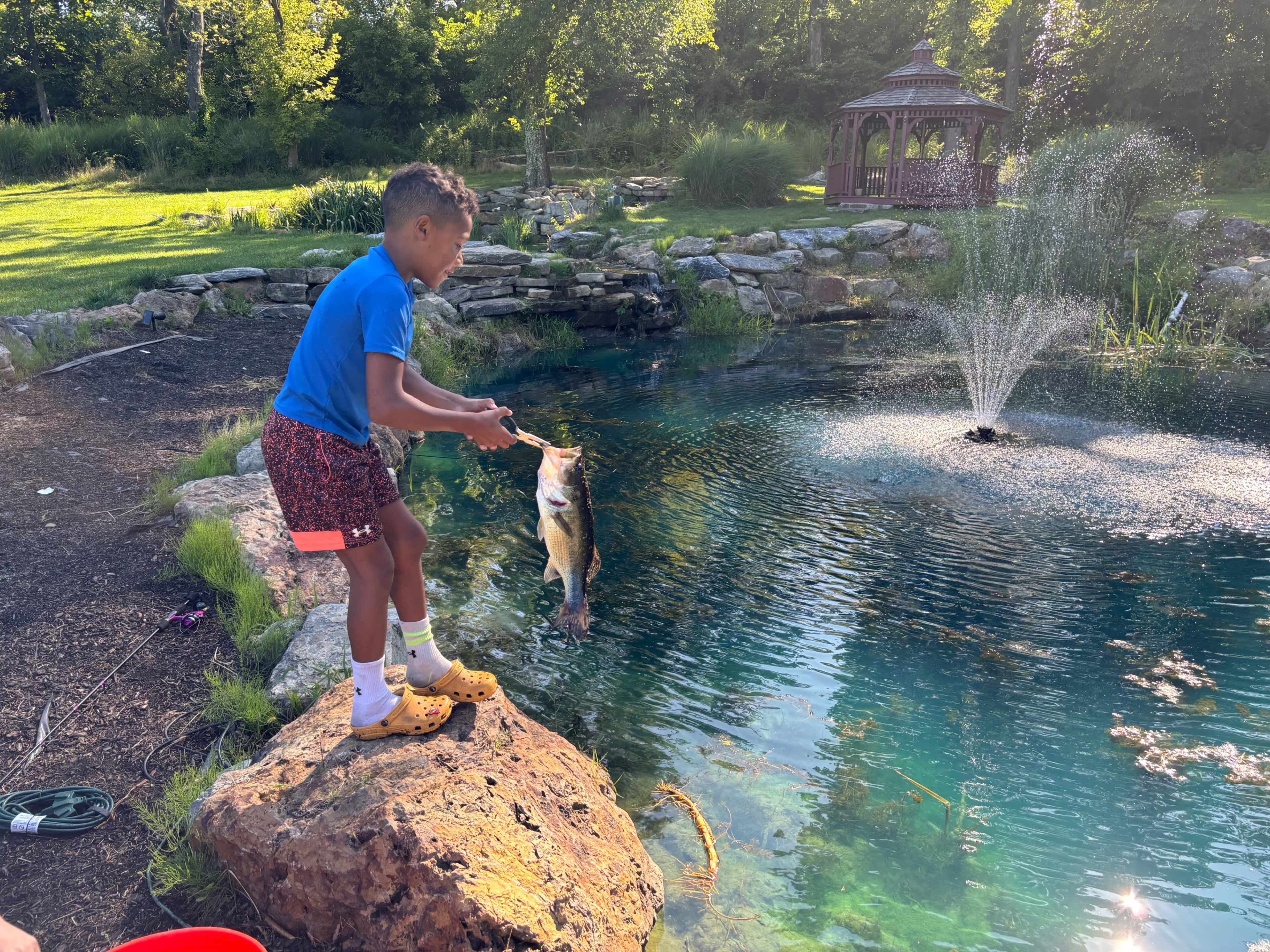 A young boy in shorts and sandals holds a large fish while standing on a rock by a pond with a fountain and a gazebo in the background.