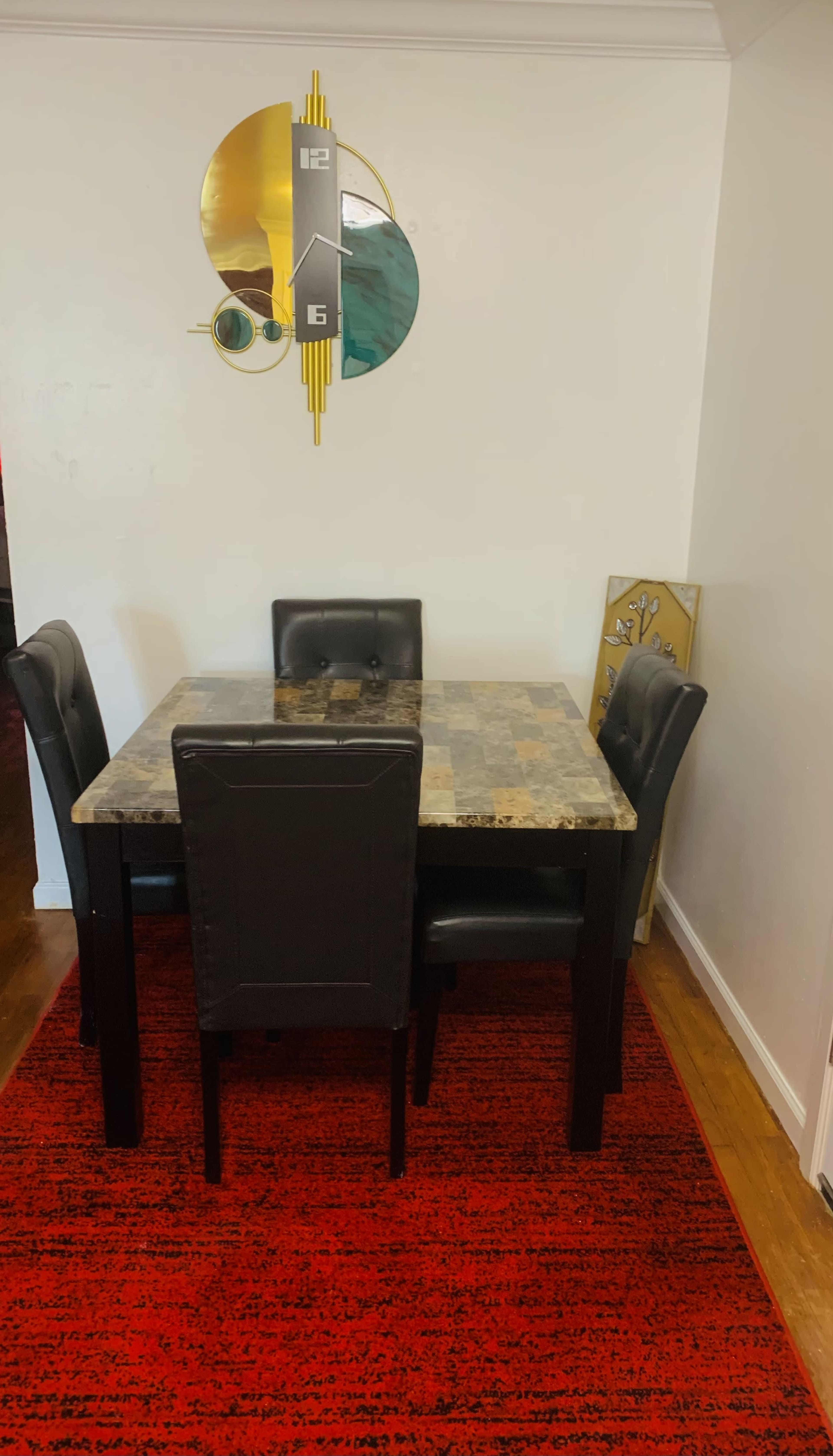 A square dining table with four black chairs is positioned on a red rug against a white wall, featuring a vibrant wall clock above it.
