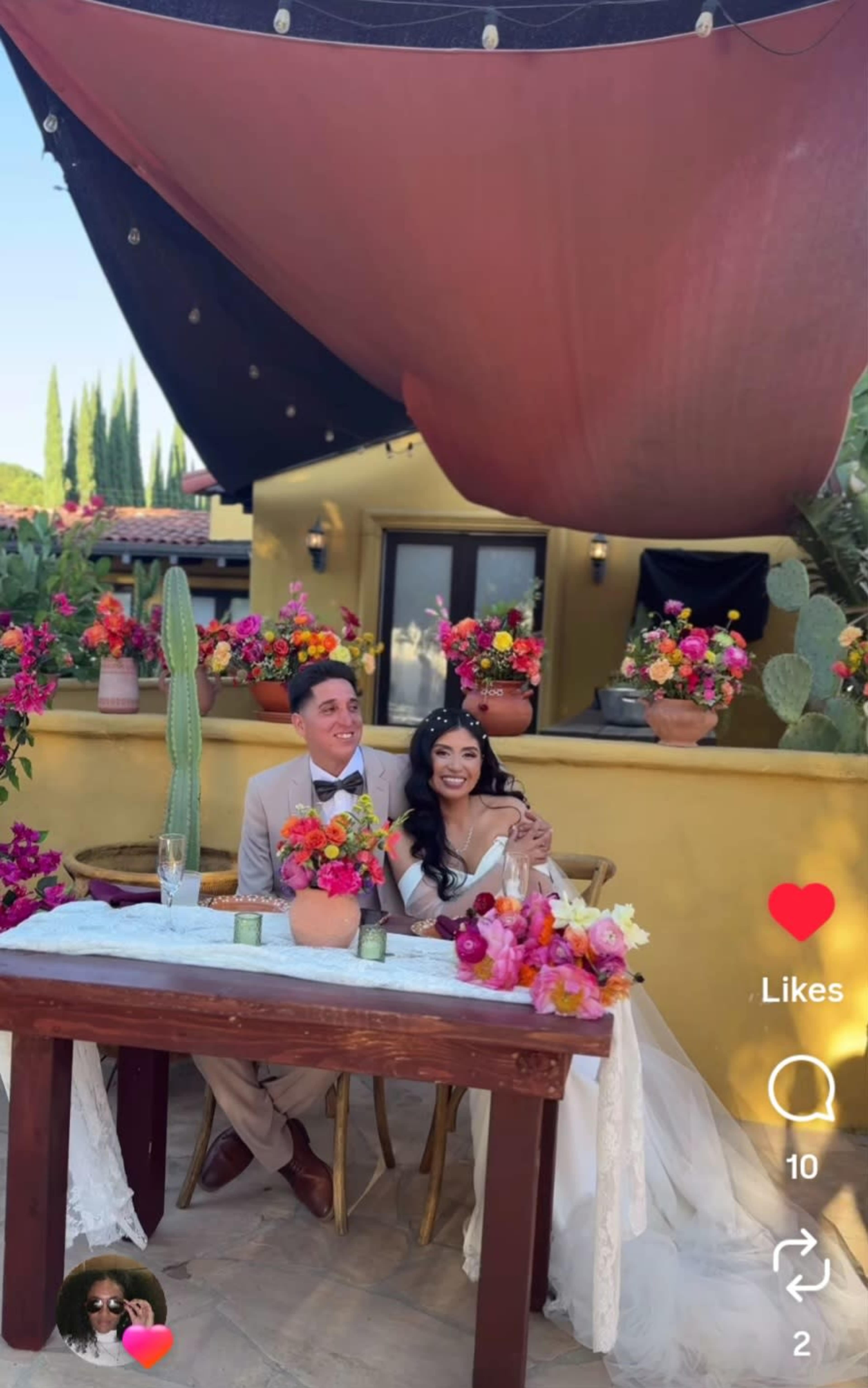A newlywed couple sits at a decorated table adorned with flowers under a canopy in an outdoor setting.