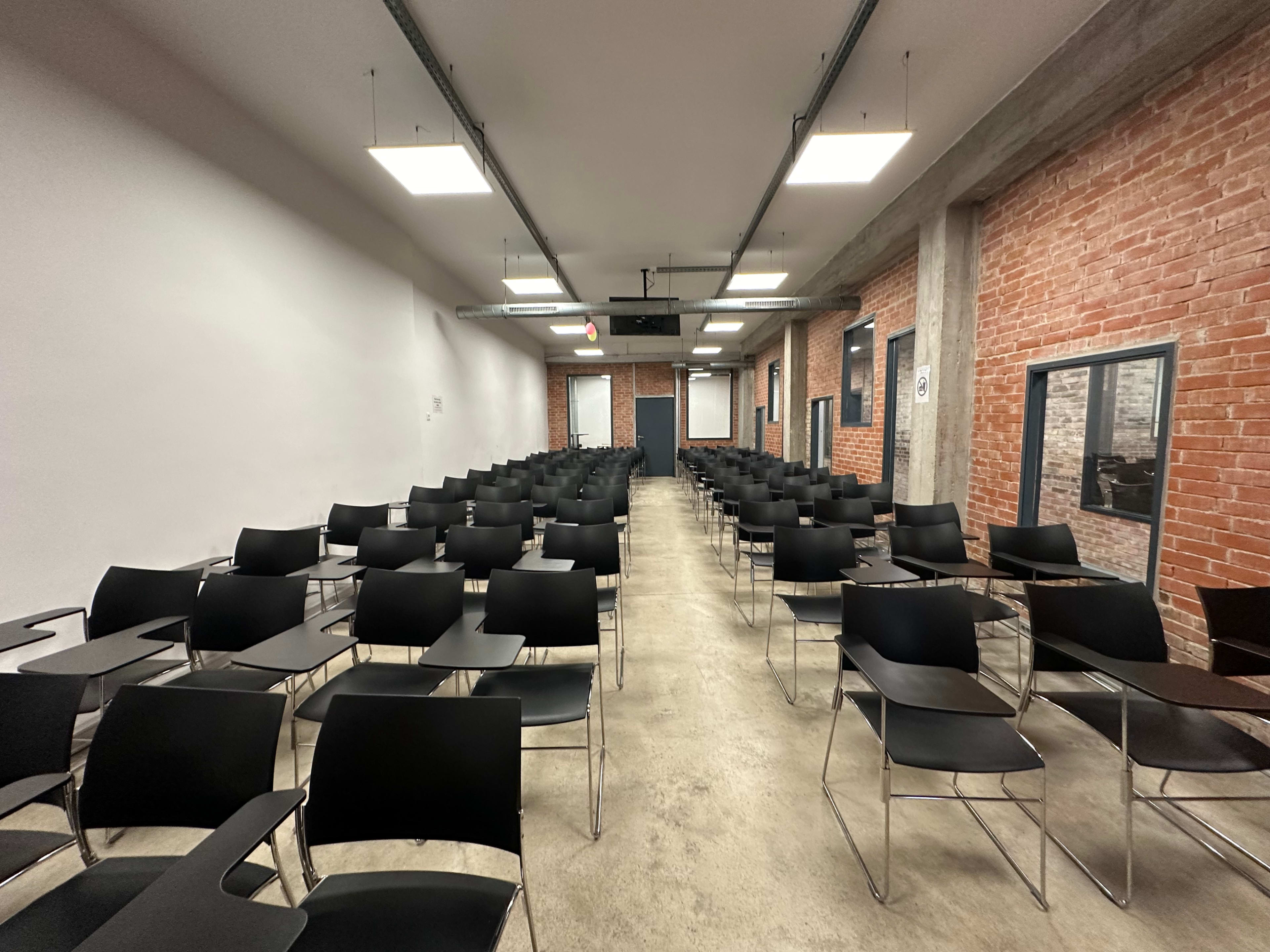 An empty classroom arranged with numerous black chairs in rows facing a front wall.