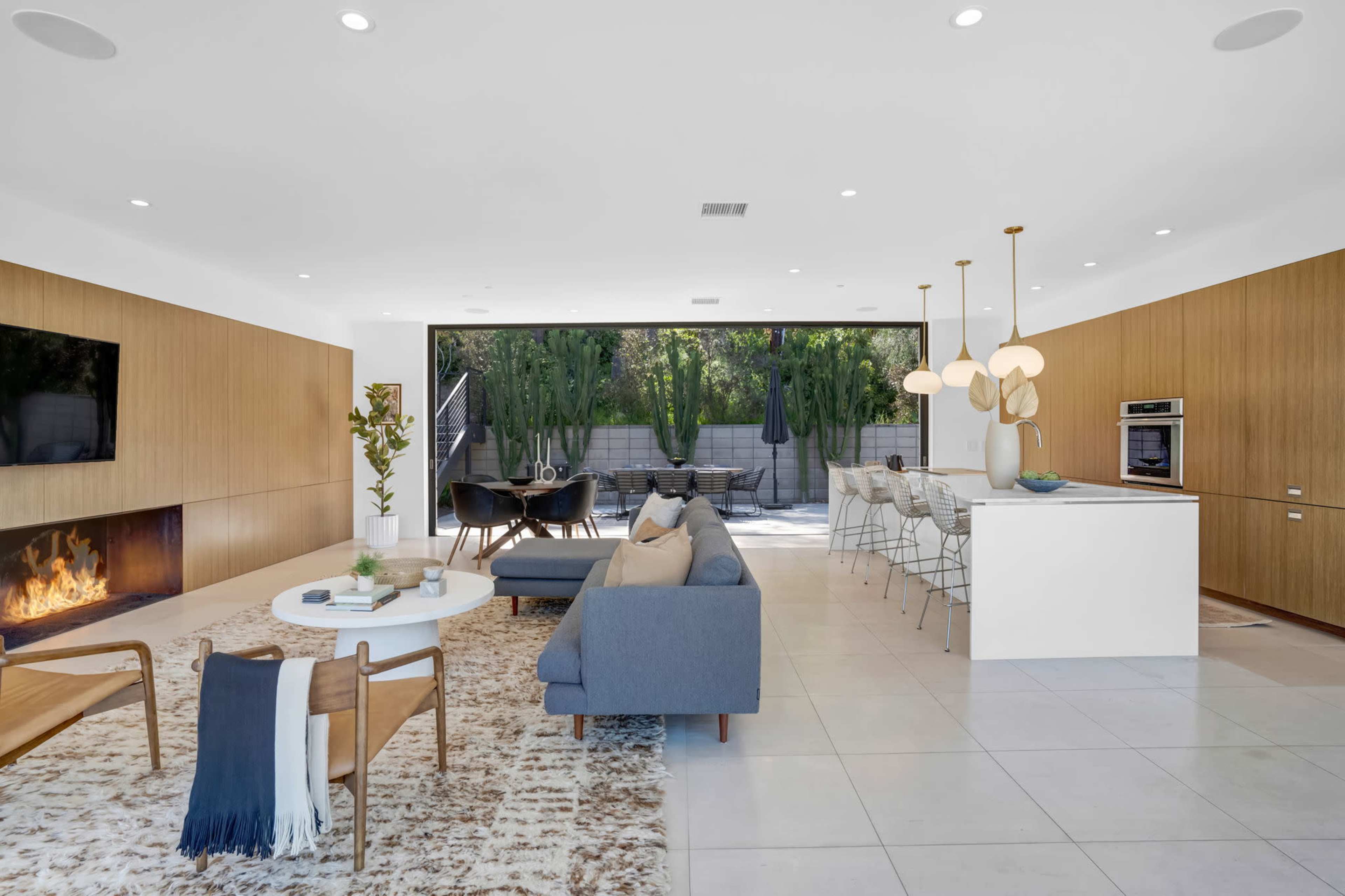 A modern living space with a fireplace, a seating area, and an open kitchen that leads to an outdoor patio surrounded by greenery.