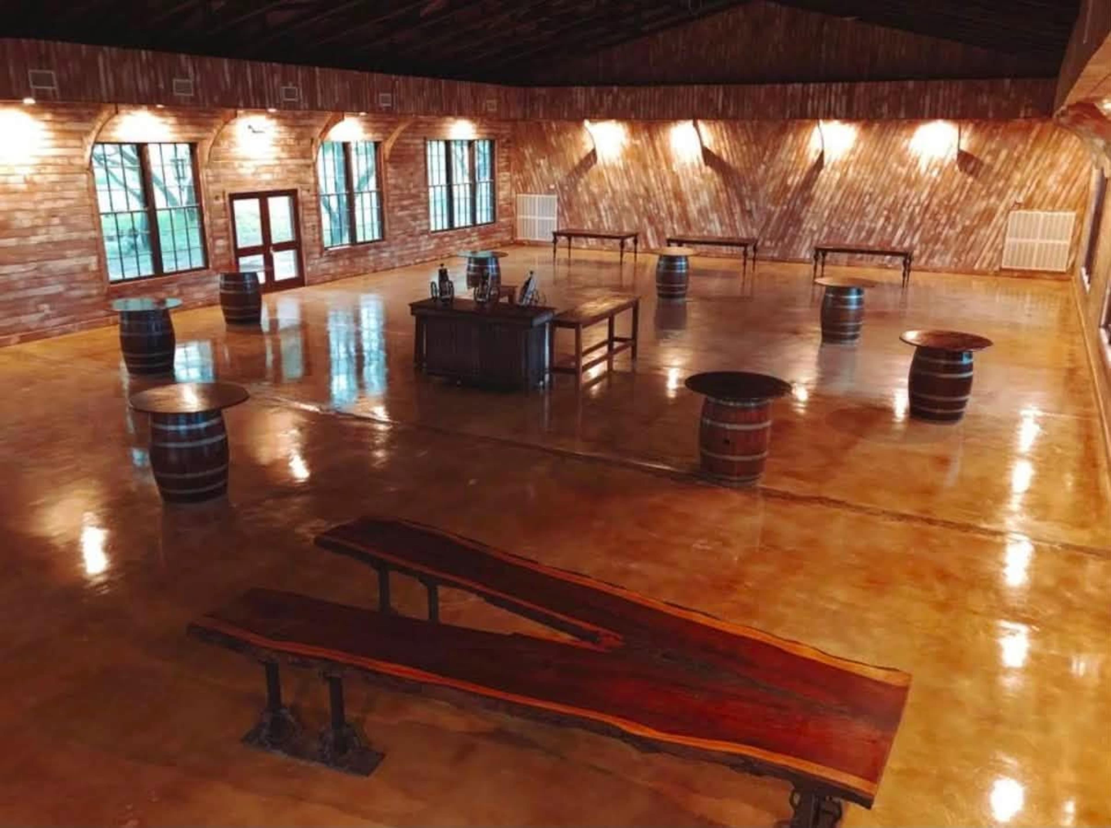 The image shows a spacious, wooden interior venue with polished concrete flooring, featuring multiple circular tables and a central rectangular table on a raised platform.
