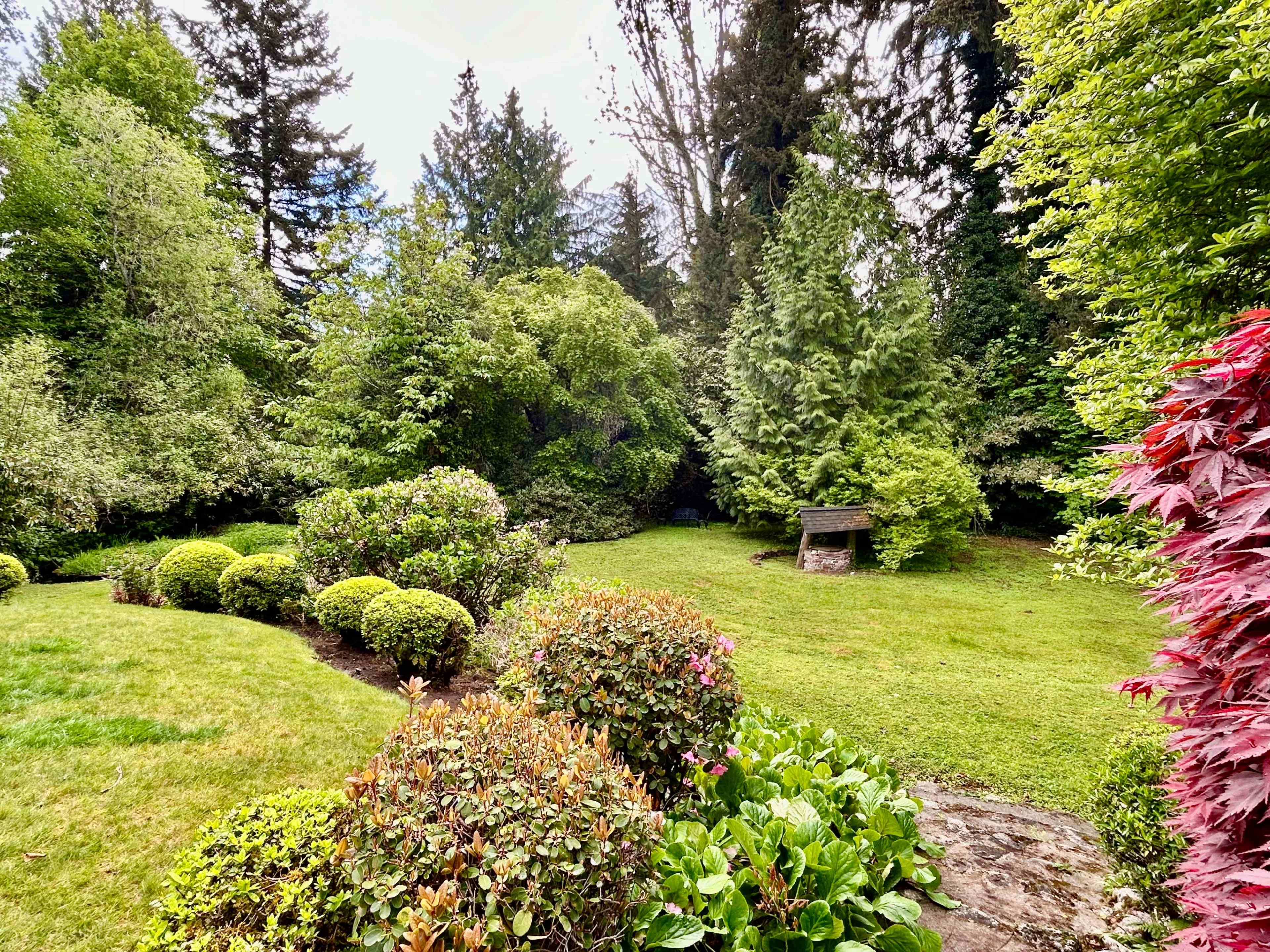 A landscaped garden features neatly trimmed bushes, vibrant greenery, and a small wooden structure in the background.