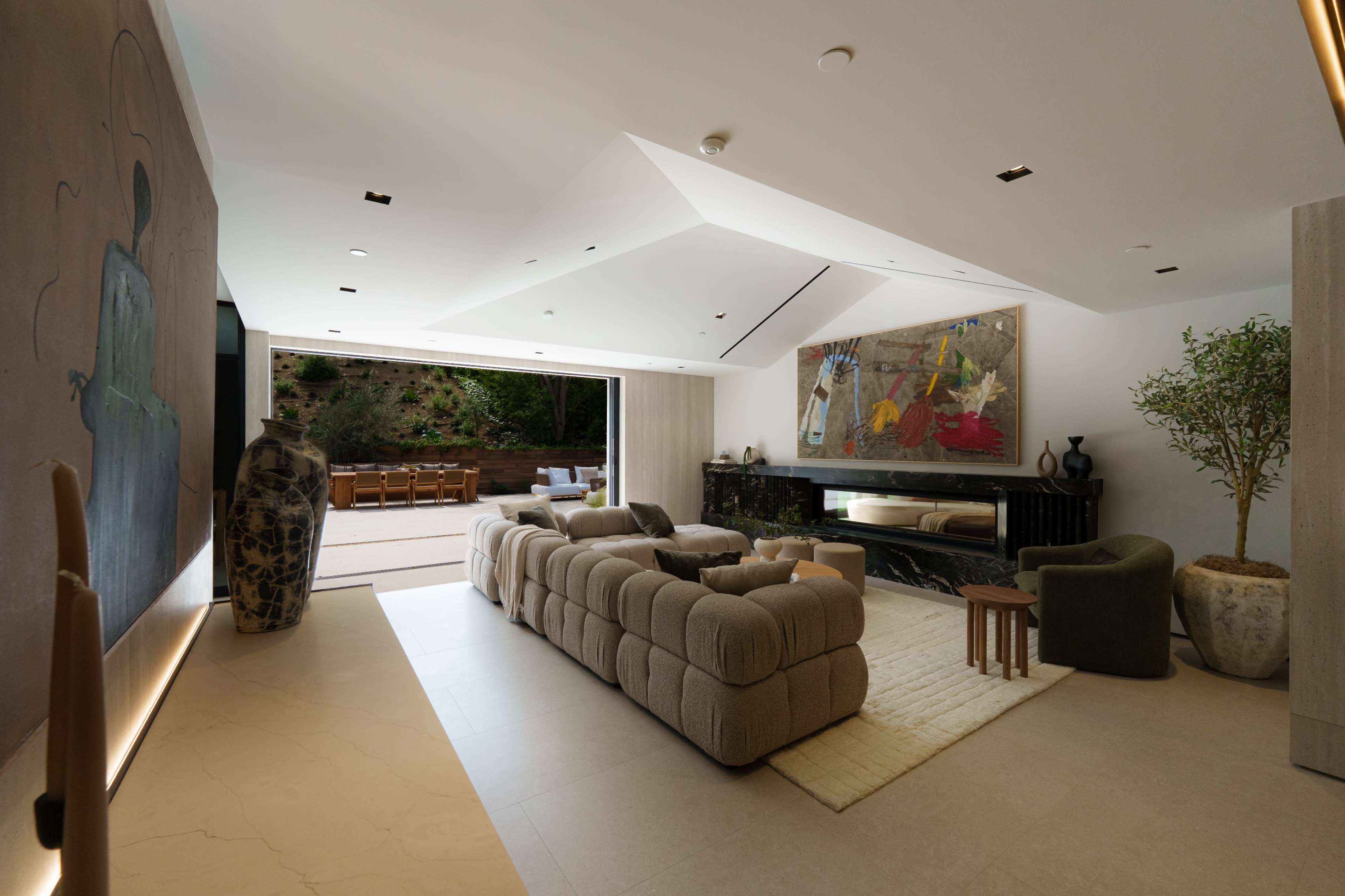The image shows a modern living room with a large sofa, abstract artwork, a fireplace, and an open door leading to an outdoor dining area.