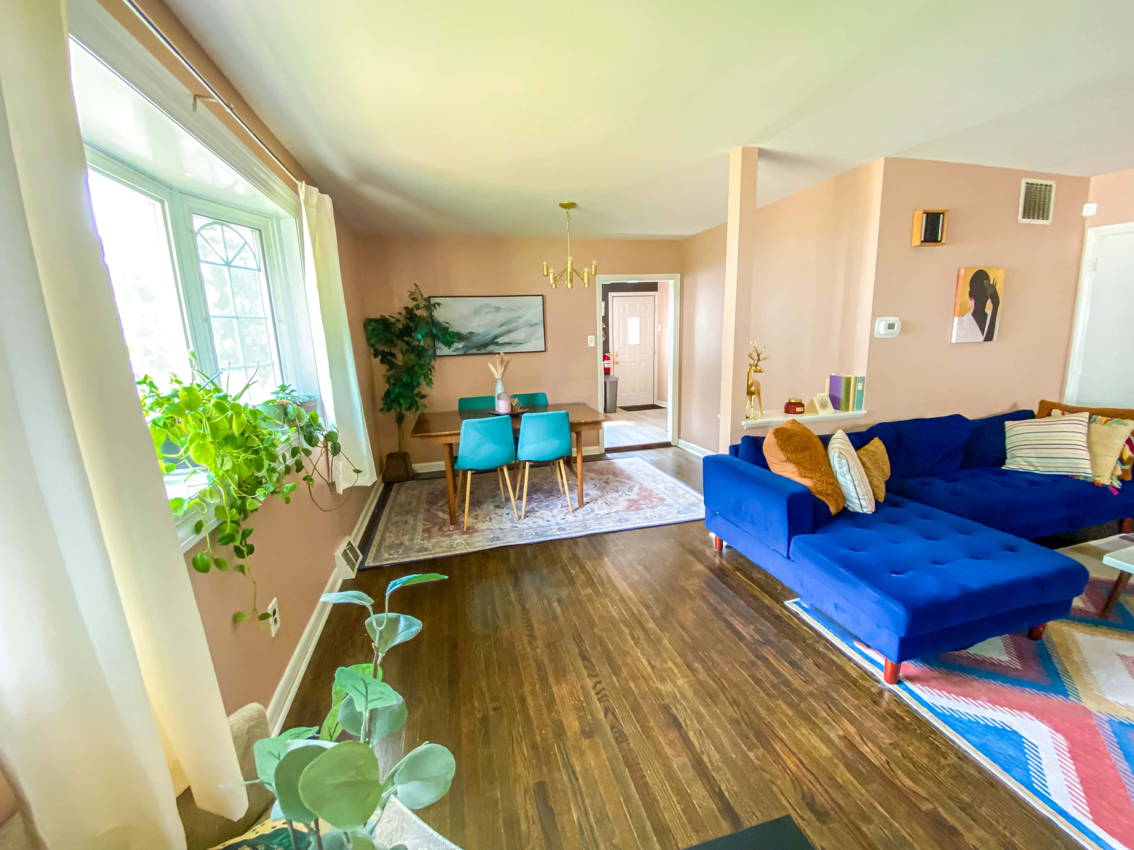 A bright living room features a blue sectional sofa, a wooden dining table, and large windows with plants near the corners.