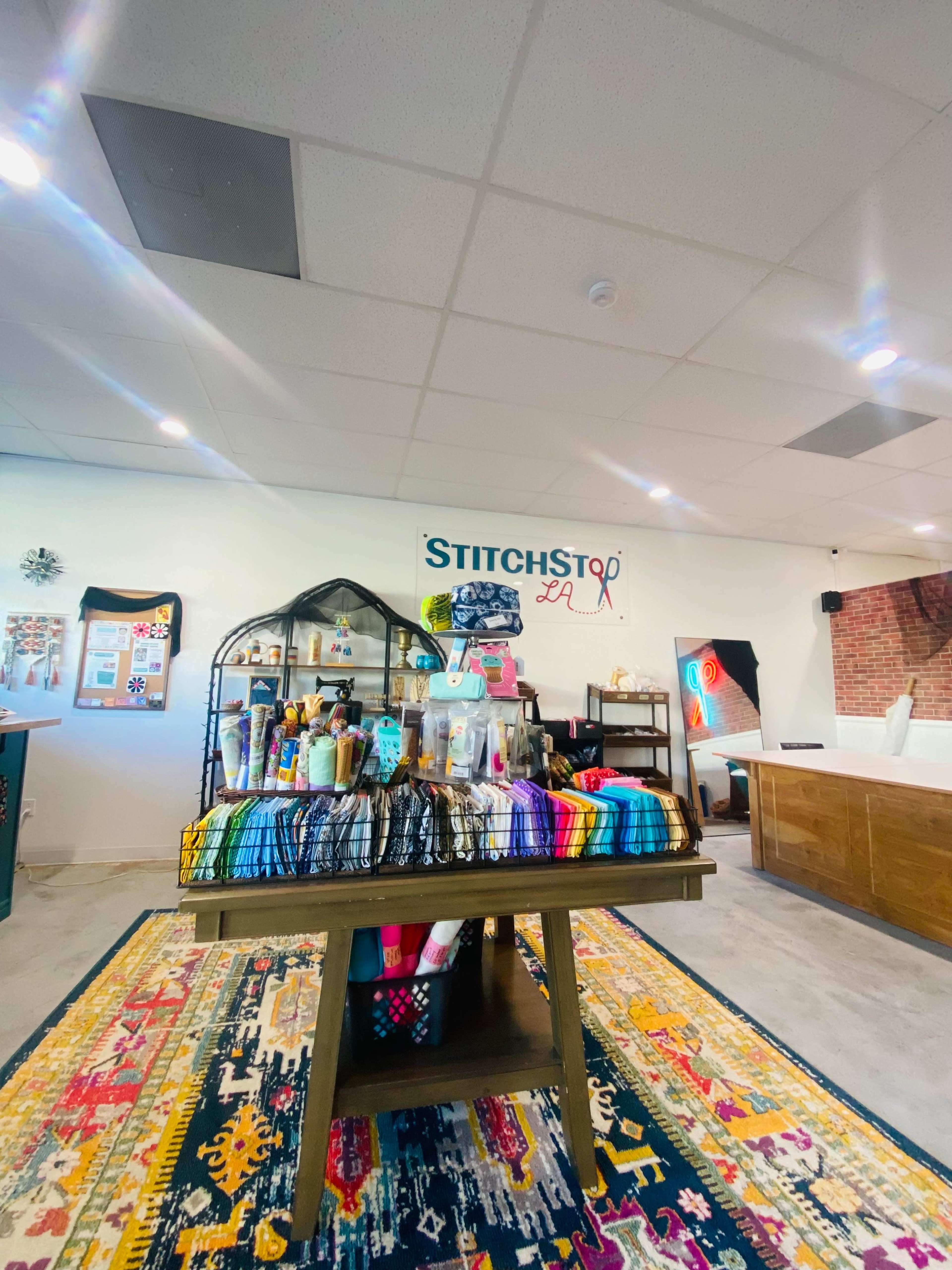 The image shows a colorful display of craft supplies and materials on a wooden table in a well-lit room decorated with various artworks and a sign reading "STITCH STOP LA."