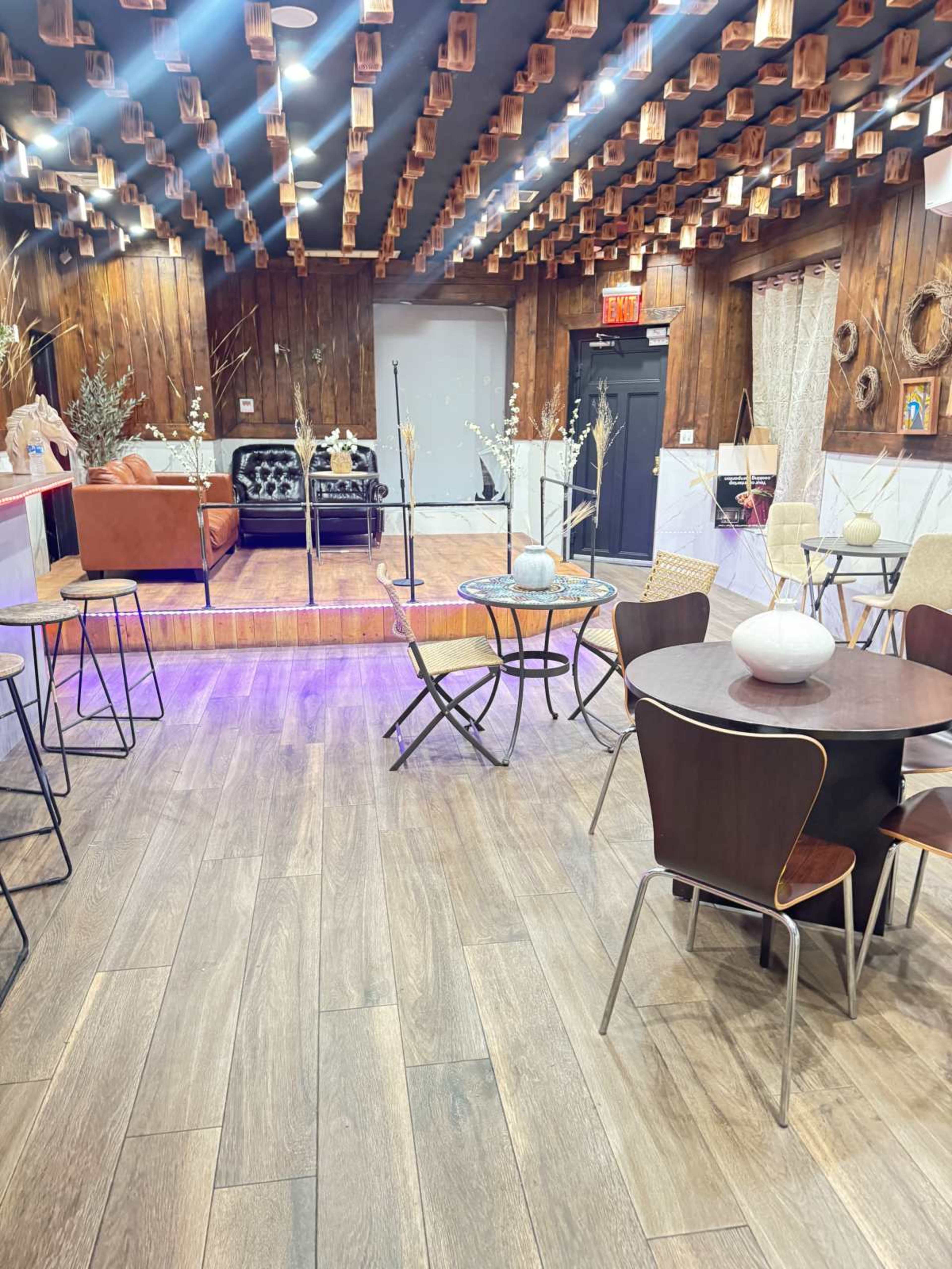 The image shows a cozy lounge area with wooden décor, seating arrangements including tables and chairs, and a small stage at the back.
