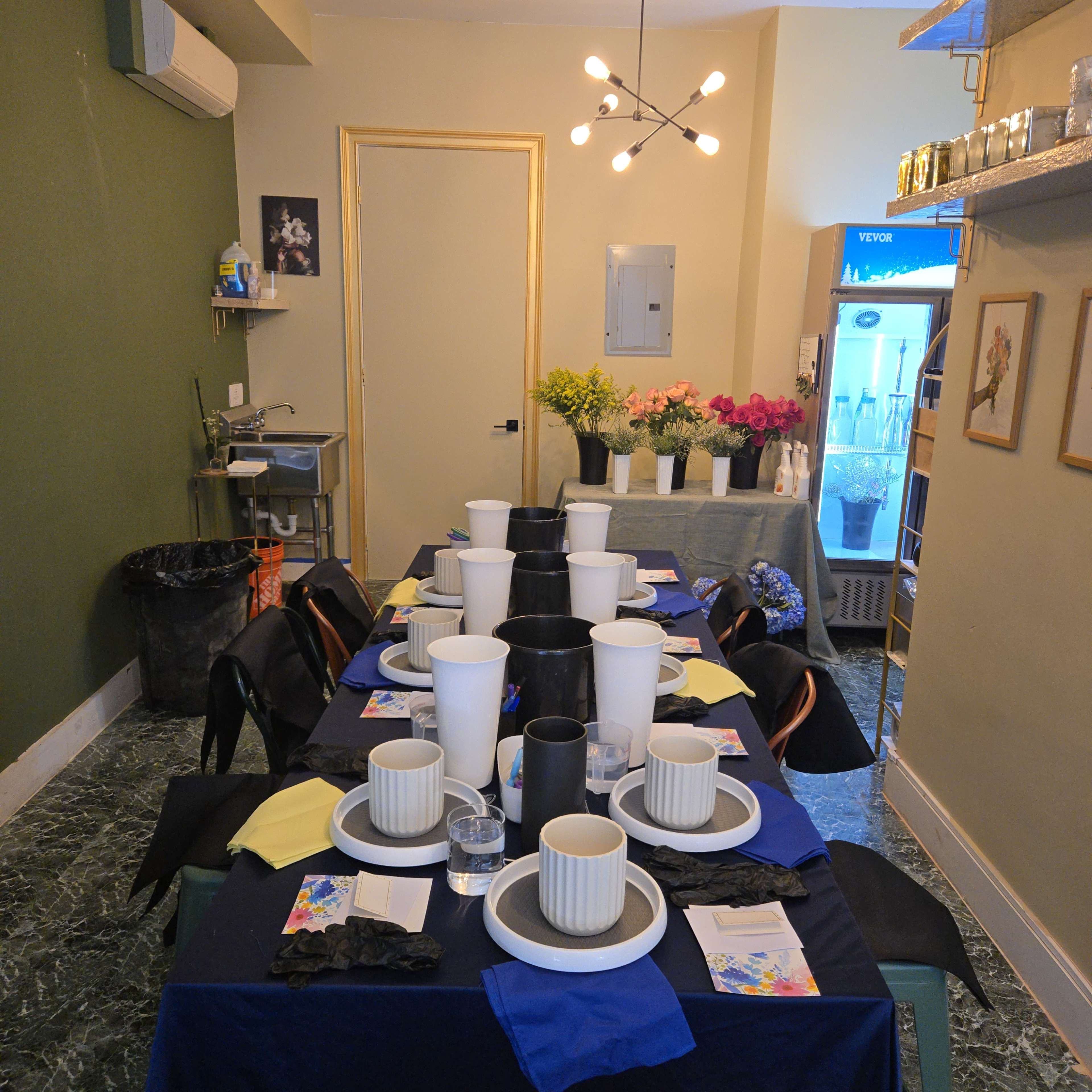 A dining table is set with plates, cups, and napkins, surrounded by a green wall, while a fridge and sink are visible in the background.