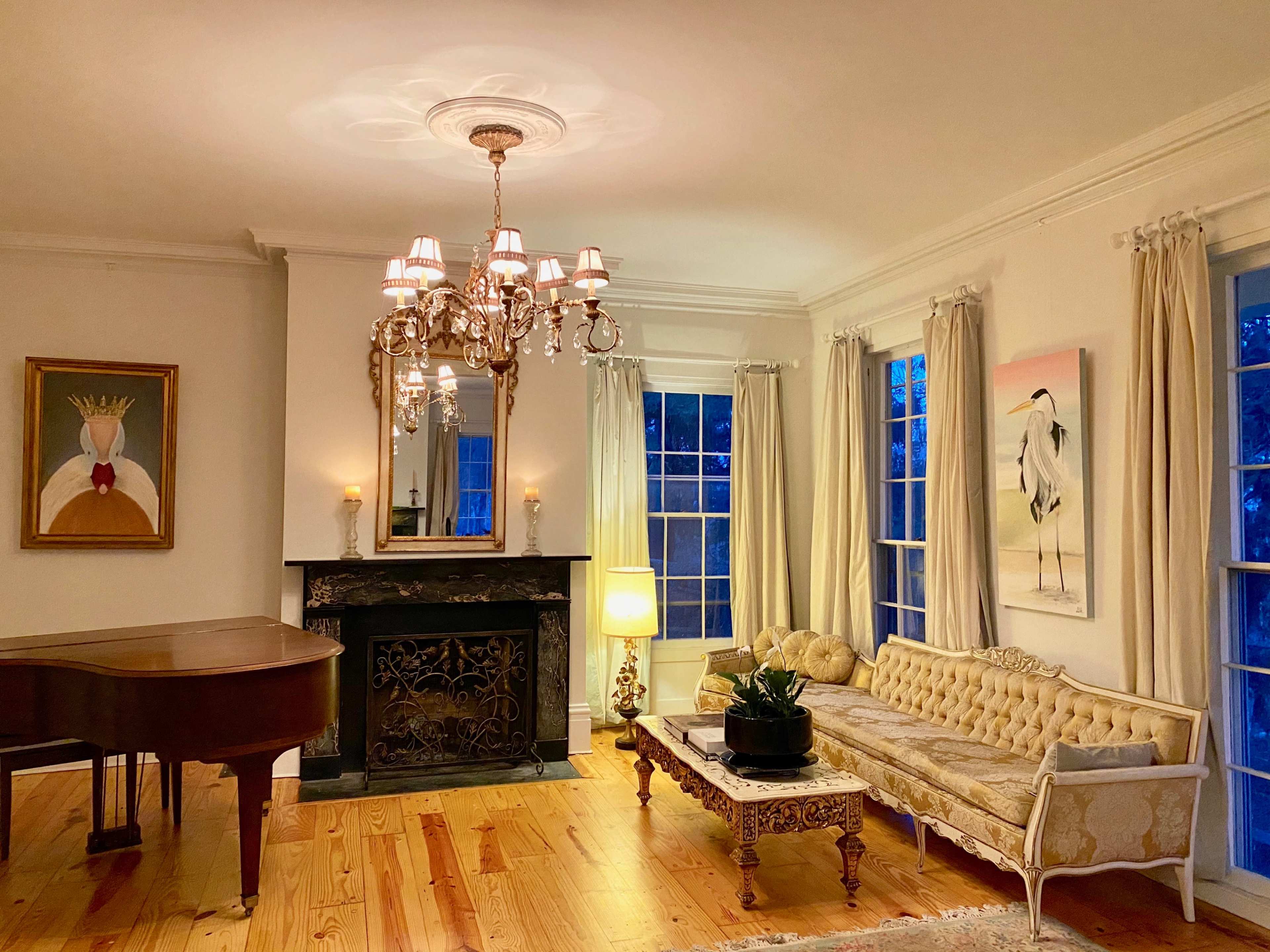A elegantly furnished living room with a chandelier, a grand piano, a floral sofa, a decorative fireplace, and large windows allowing natural light.