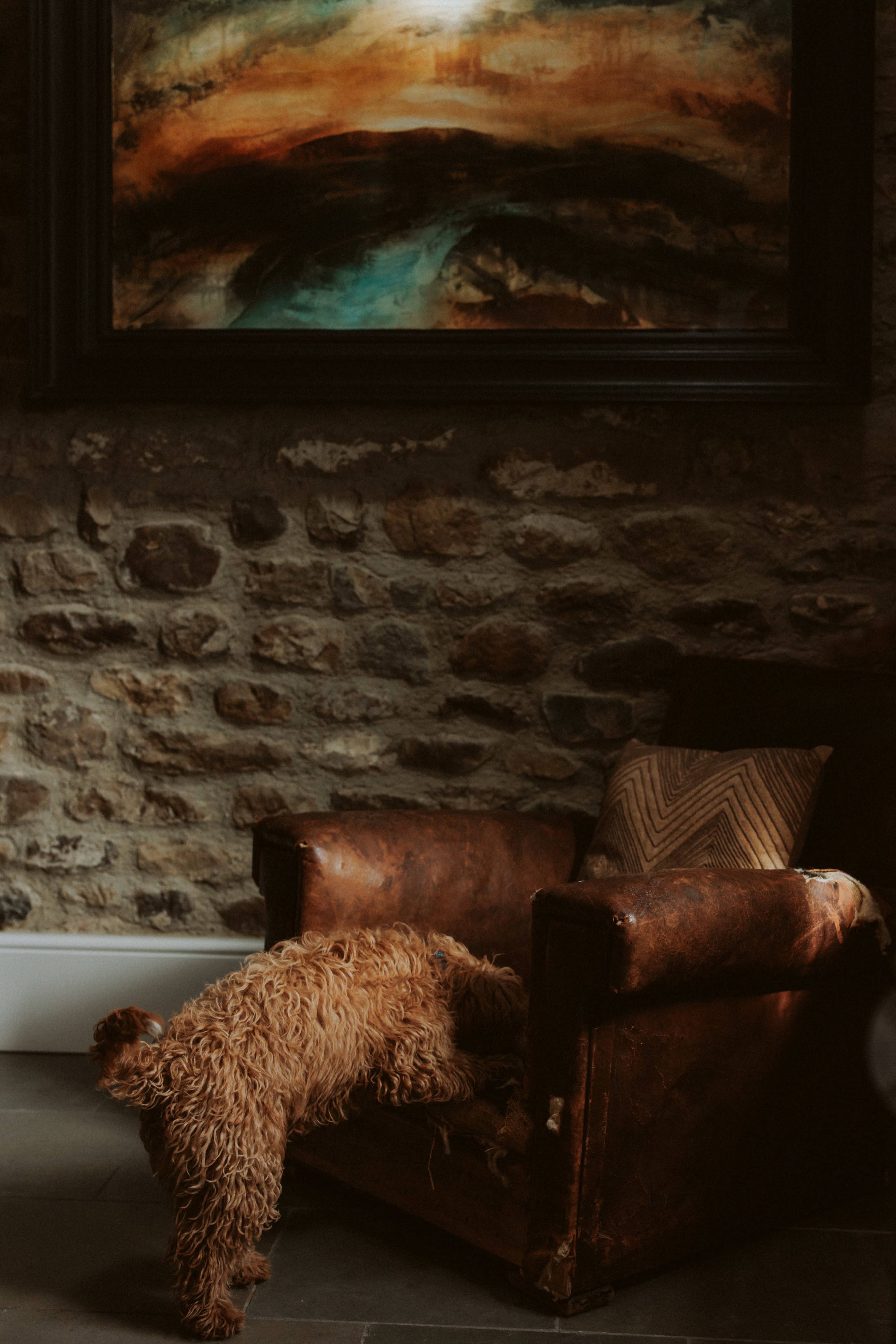 A dog is climbing onto a worn leather armchair in a room with stone walls and a large painting above it.