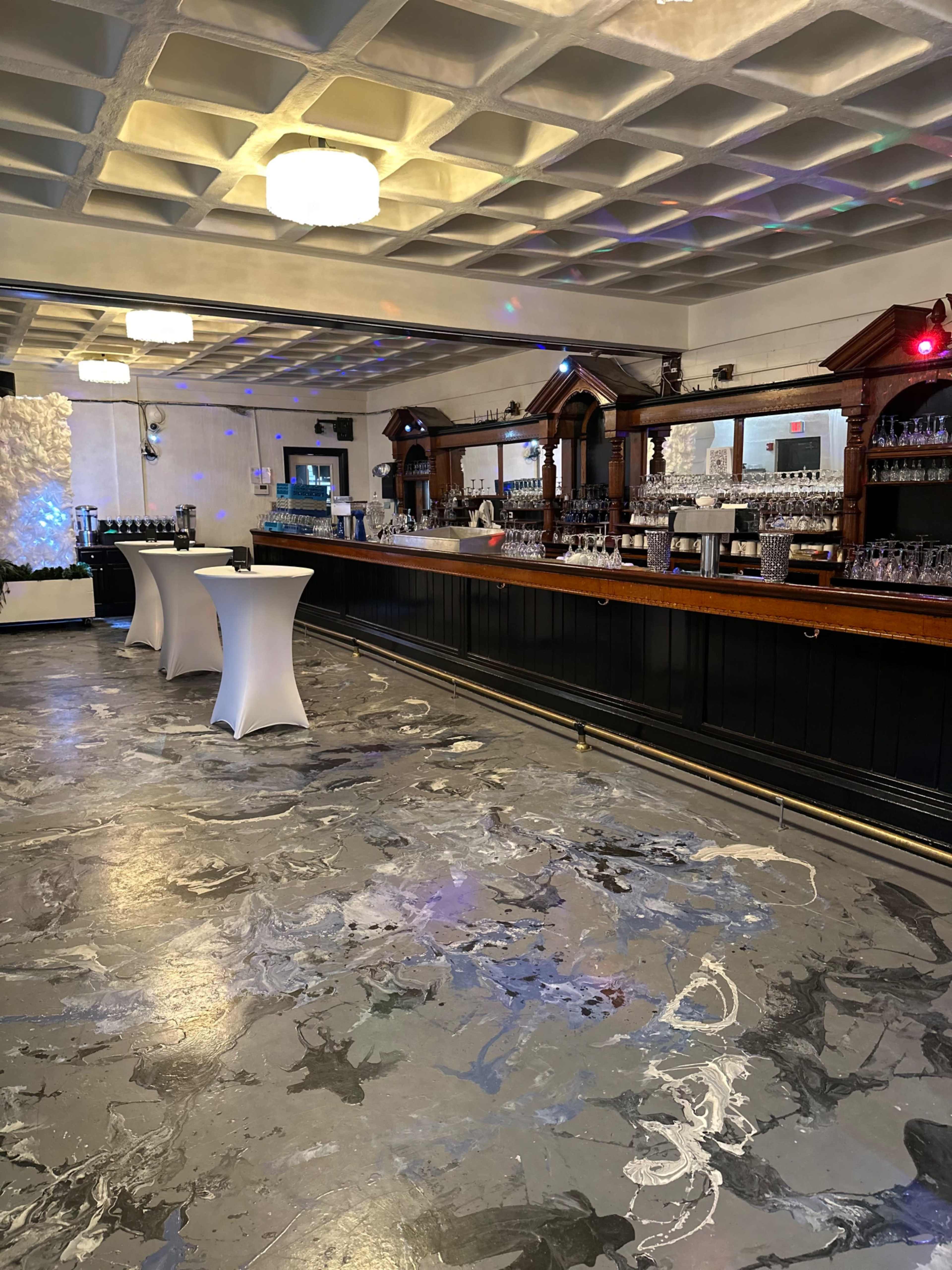 The interior of a bar features a long wooden counter lined with glasses, a marble-patterned floor, and minimalistic high tables.