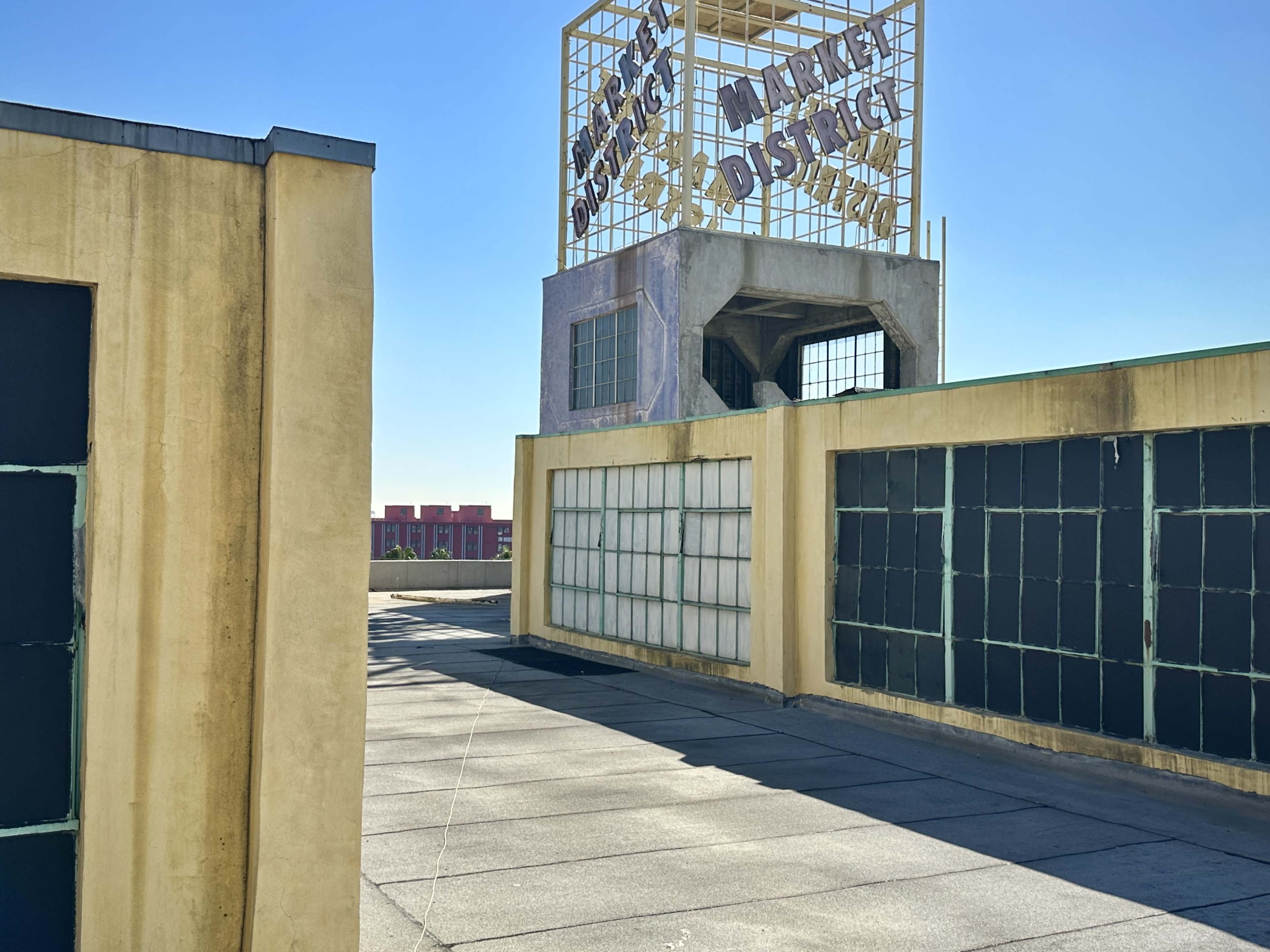 An elevated view shows a rooftop walkway with large windows and a prominent sign reading "Market District" in the background under a clear blue sky.