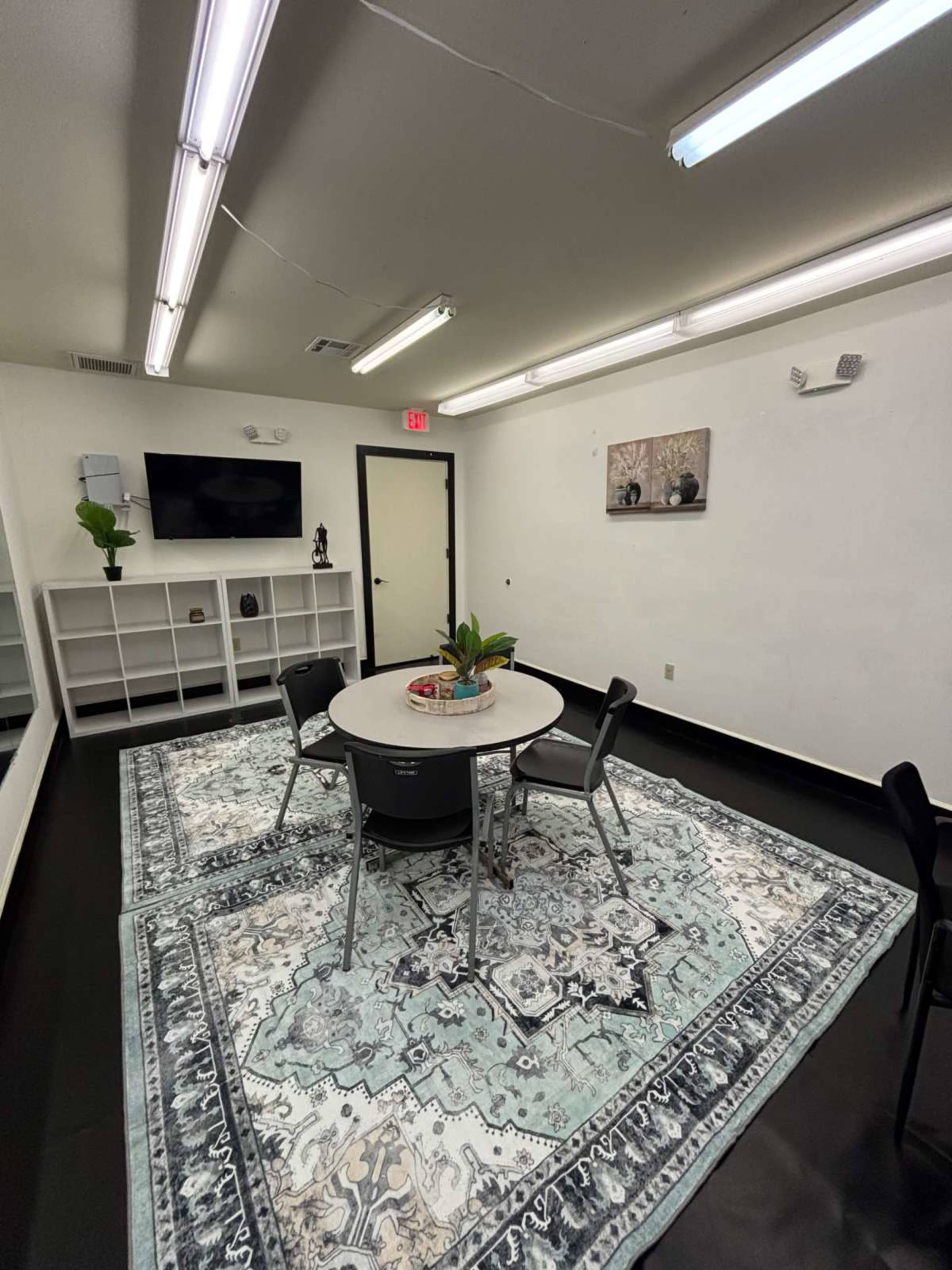 The image shows a bright room featuring a round table surrounded by four chairs, a large patterned rug, a television on the wall, and shelves with decor.