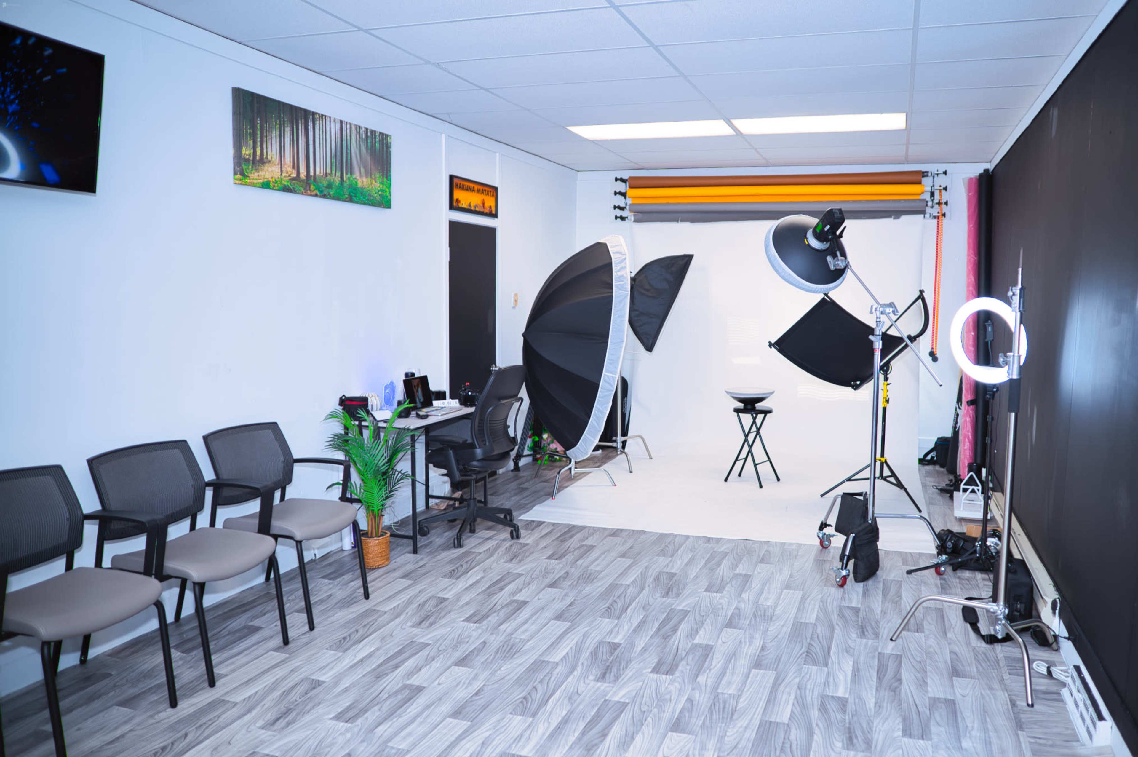 A photography studio with a waiting area featuring black chairs and a backdrop setup including softboxes and a ring light on a white backdrop.