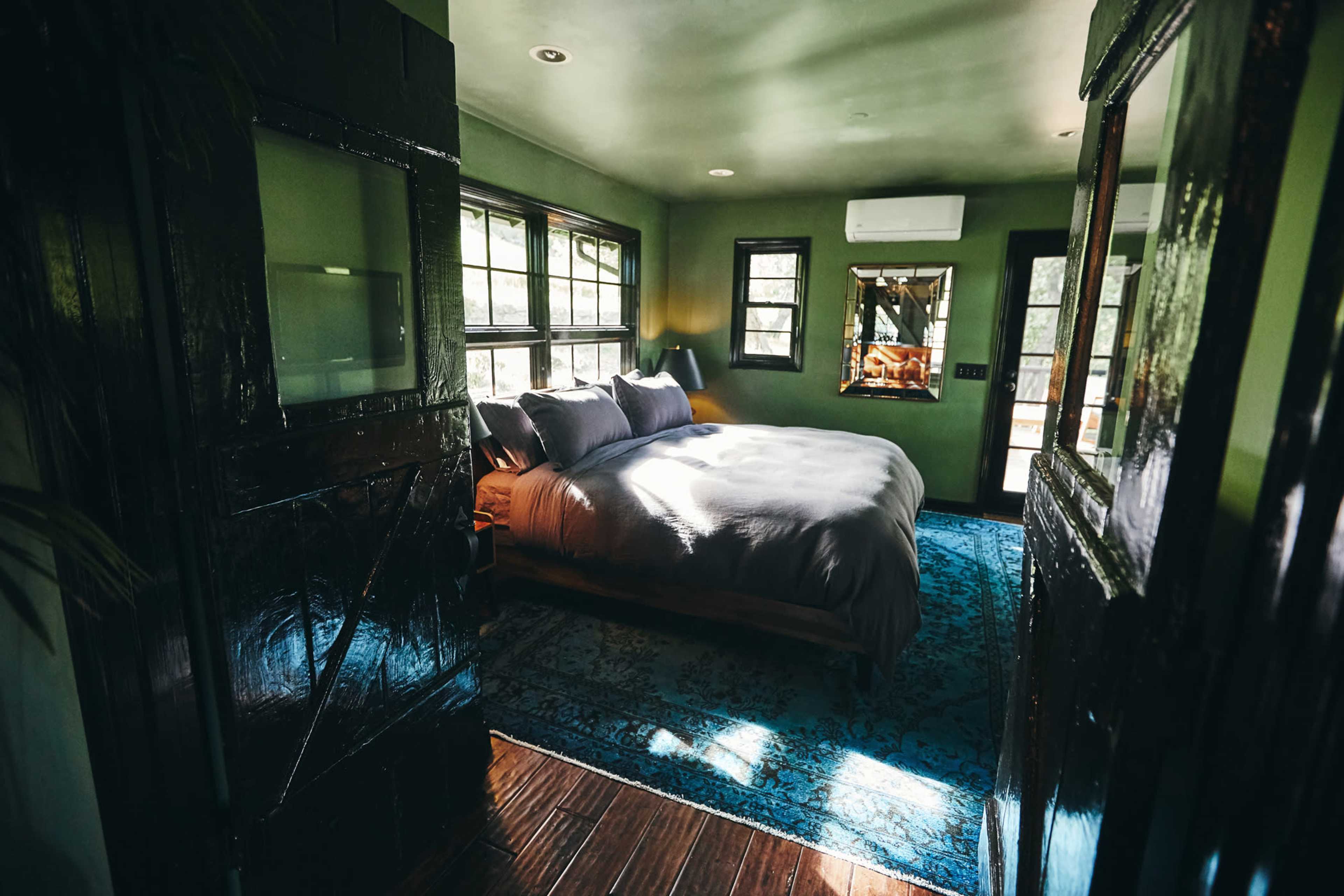 The image shows a softly lit bedroom with a large bed, green walls, and a blue area rug.