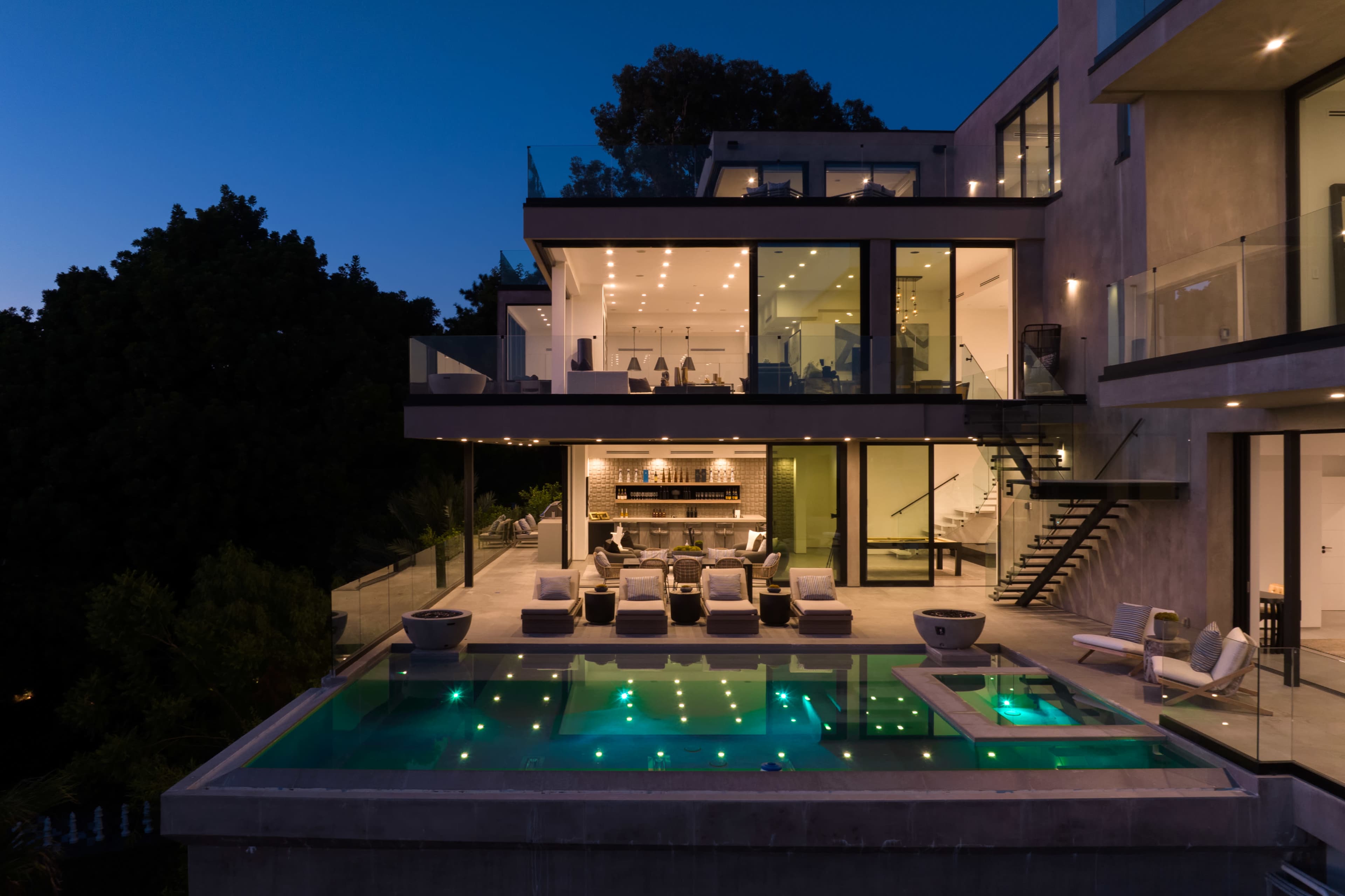 A modern two-story house features large glass windows, an outdoor pool with illuminated water, and a spacious patio area furnished with lounge chairs.