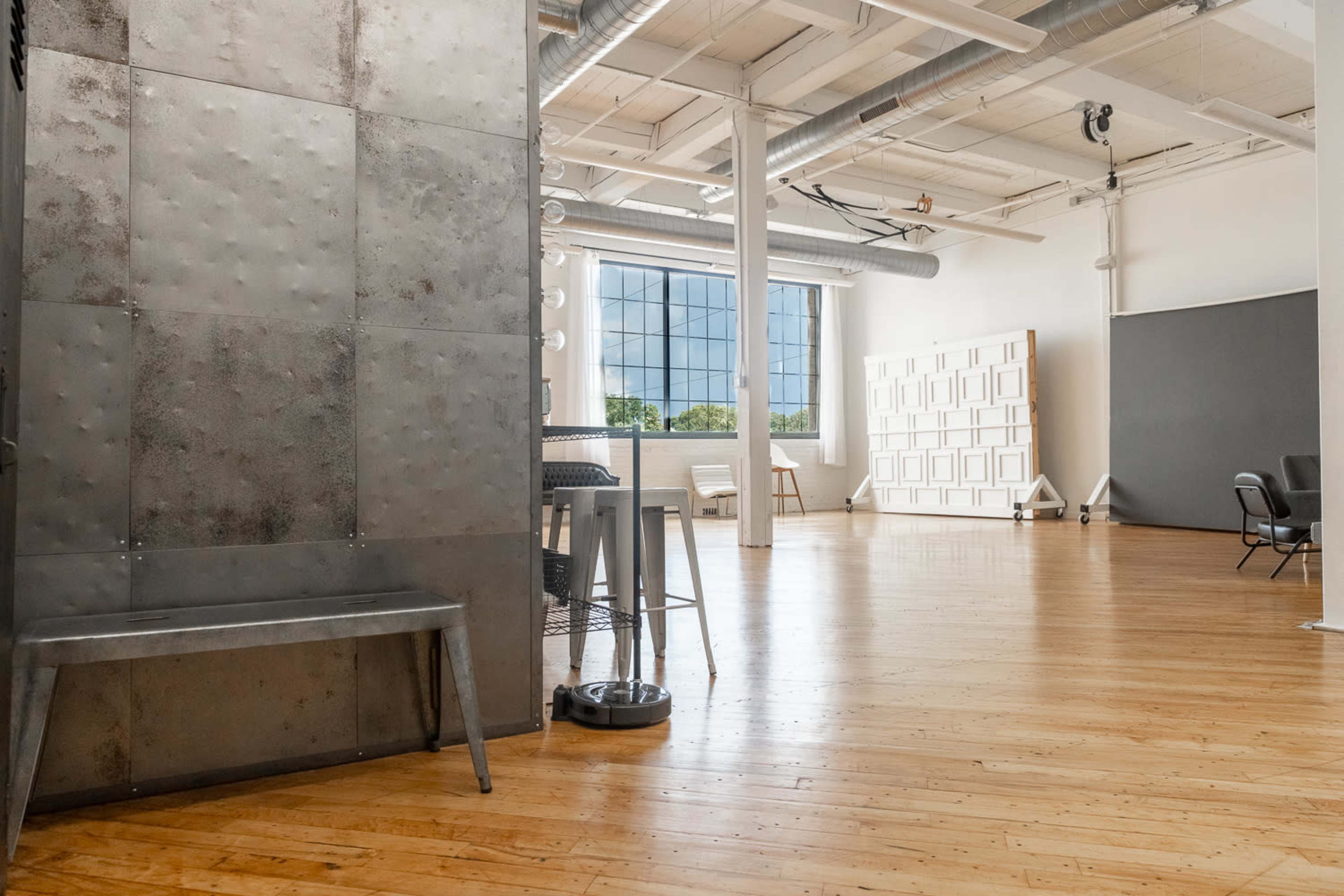 The image shows a spacious, modern studio with large windows, sleek wooden flooring, and a mix of metal and wooden furnishings.