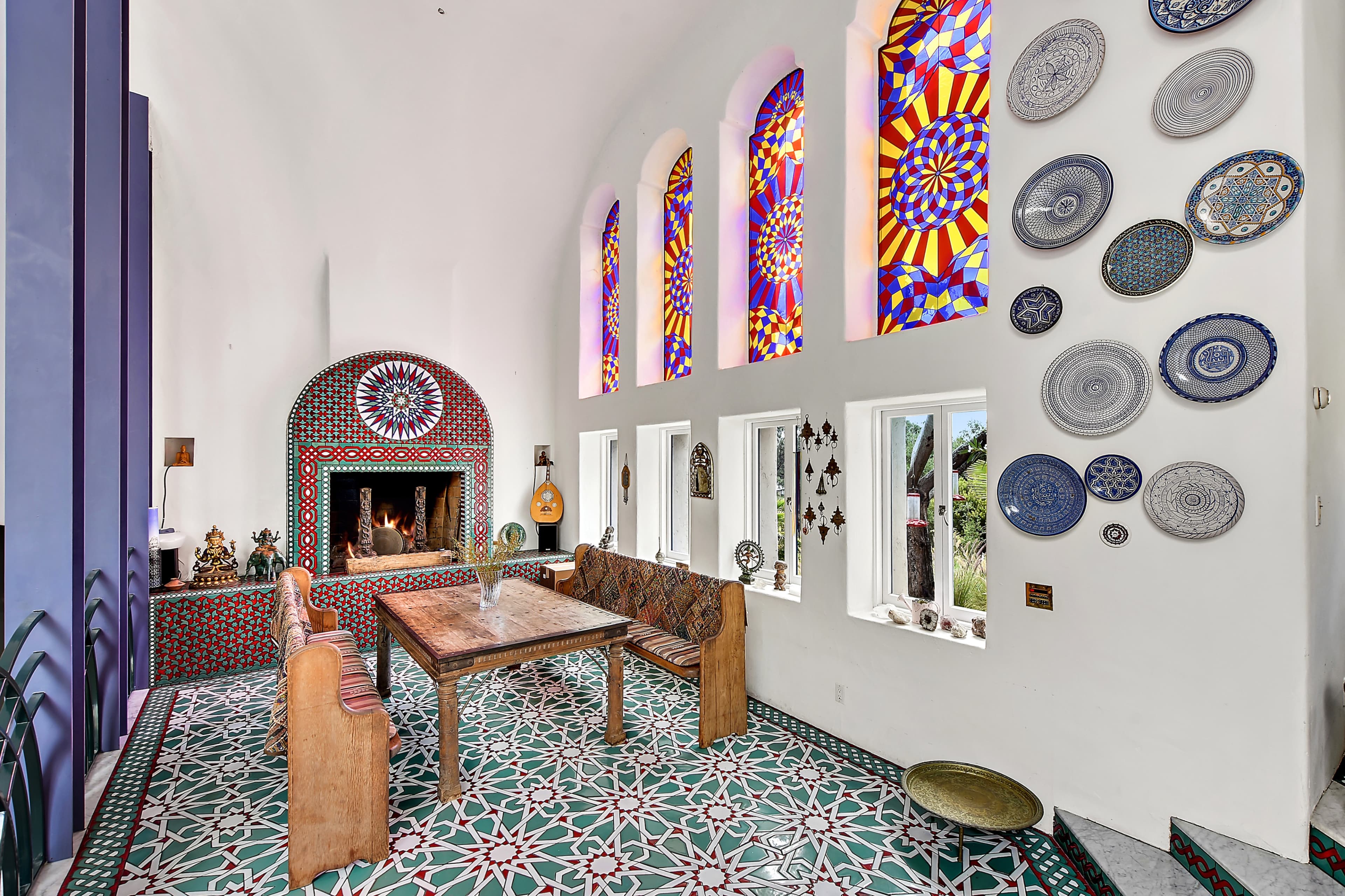 The image depicts a bright room with colorful stained glass windows, a decorative fireplace, and a table surrounded by patterned seating.