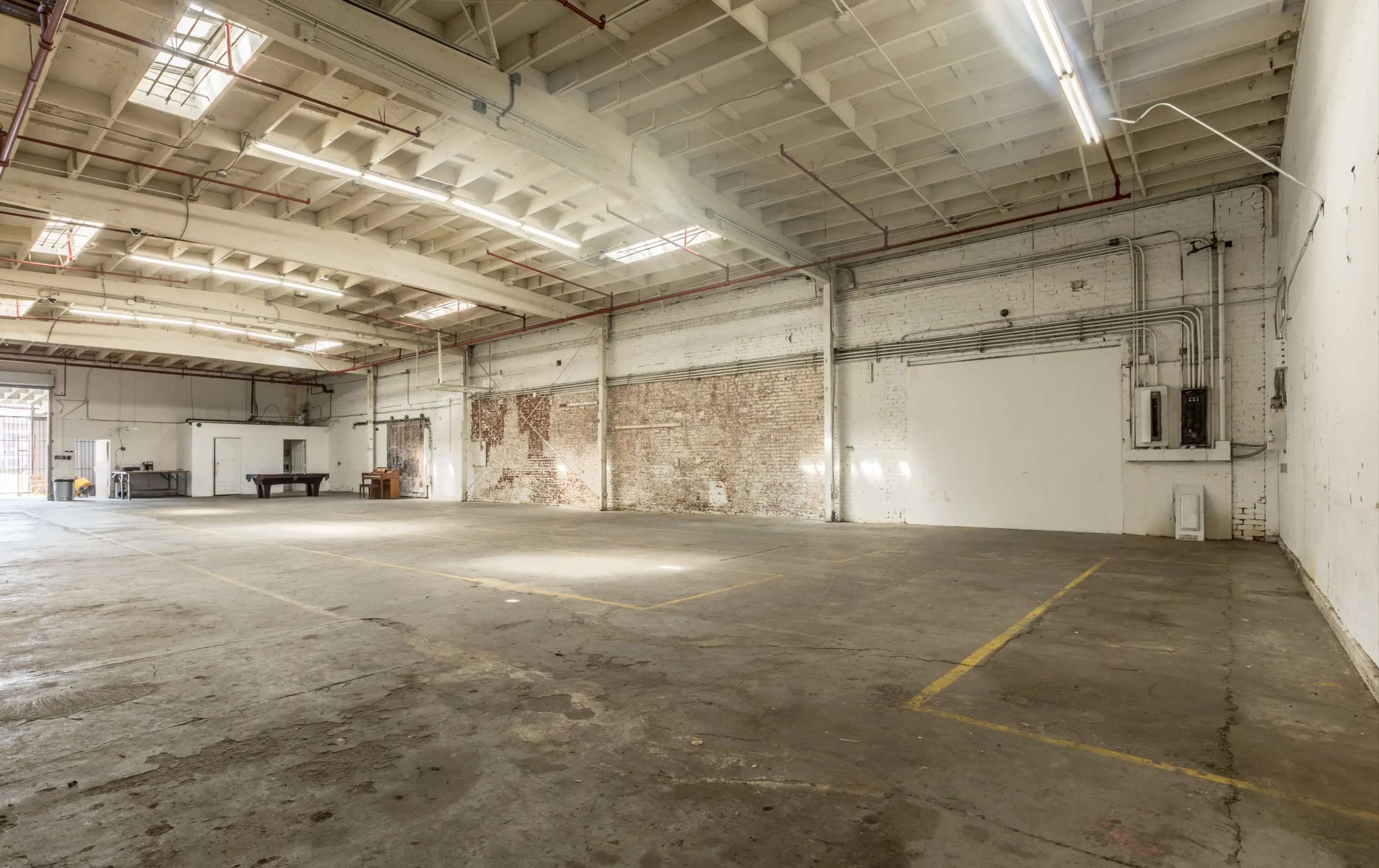 The image shows a large, empty industrial space with exposed brick walls and a concrete floor, illuminated by natural light coming through windows.