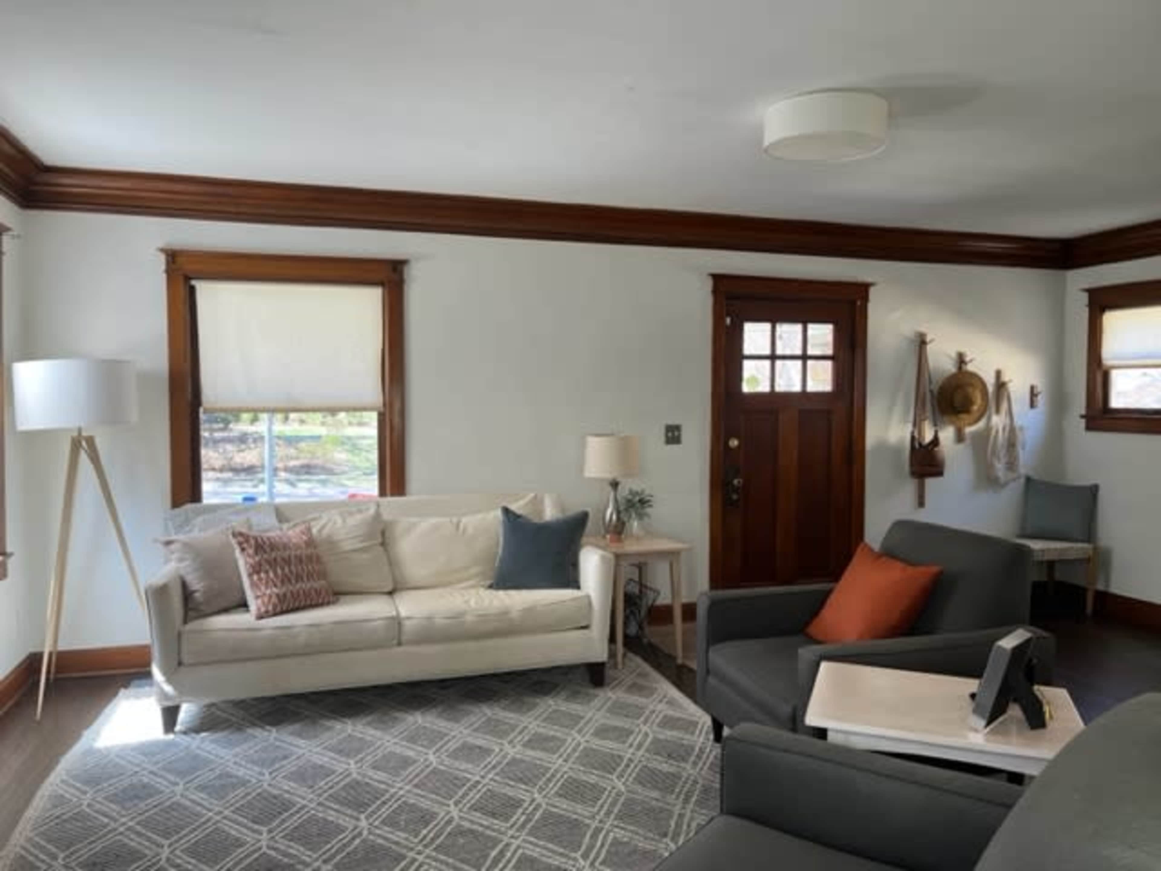 The image shows a living room with a white couch, two armchairs, a lamp, and a front door, all surrounded by light-colored walls and wood trim.