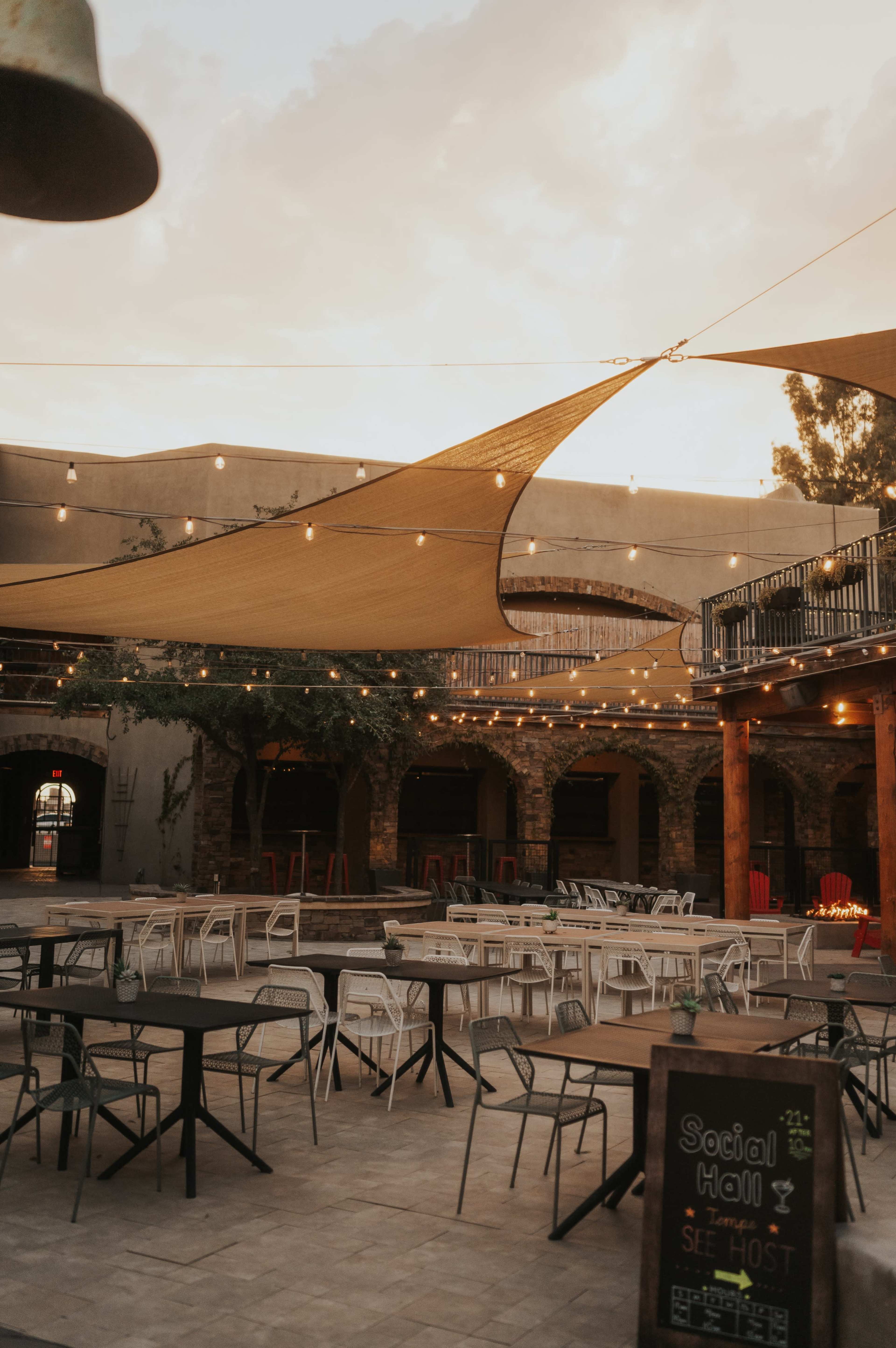 The image shows an outdoor event space with string lights, shaded seating areas, and a mix of tables arranged on a paved surface.