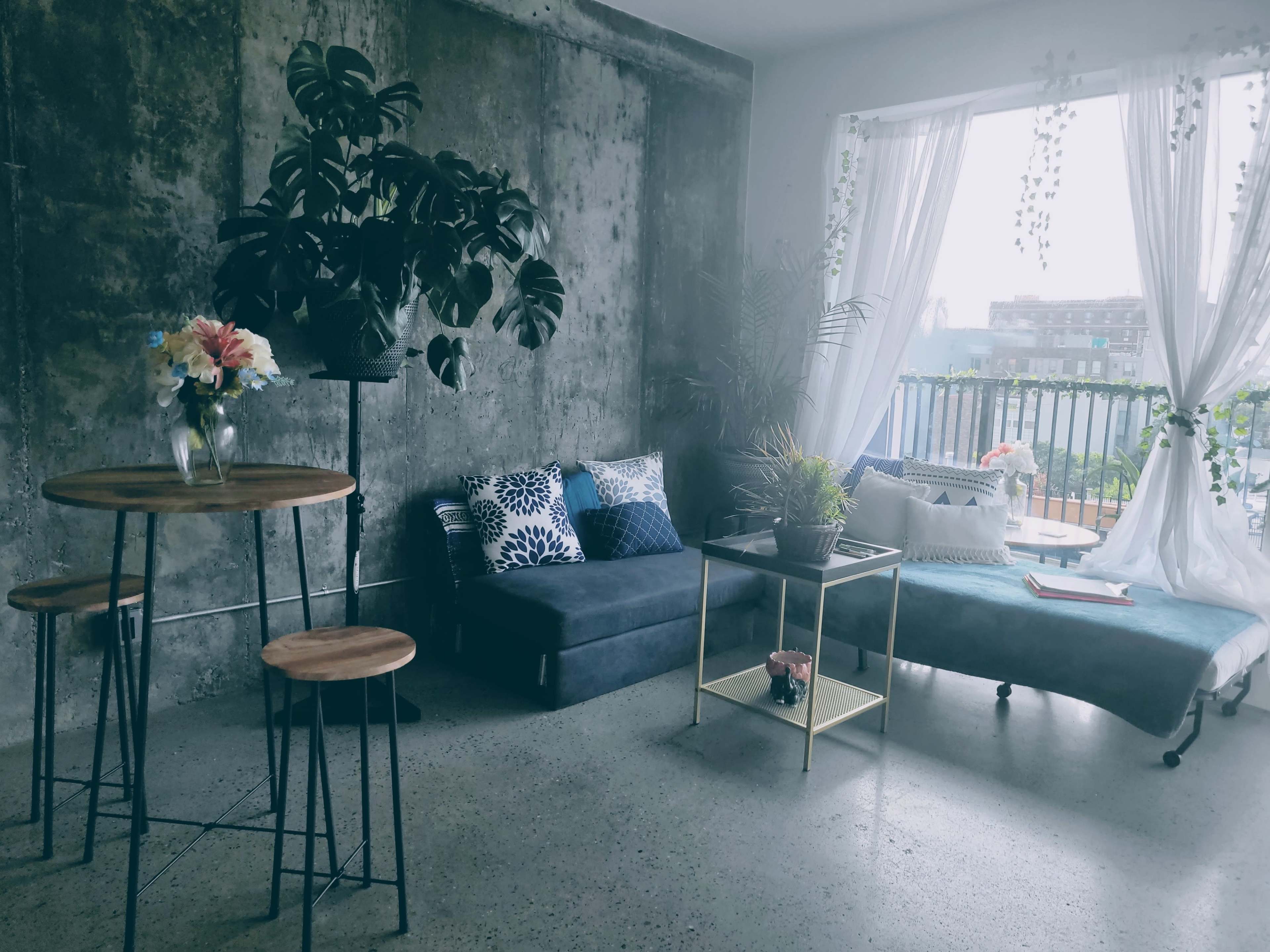 A modern living space with a concrete wall, a small round wooden table with stools, a blue sofa with decorative pillows, and large windows draped in sheer curtains.