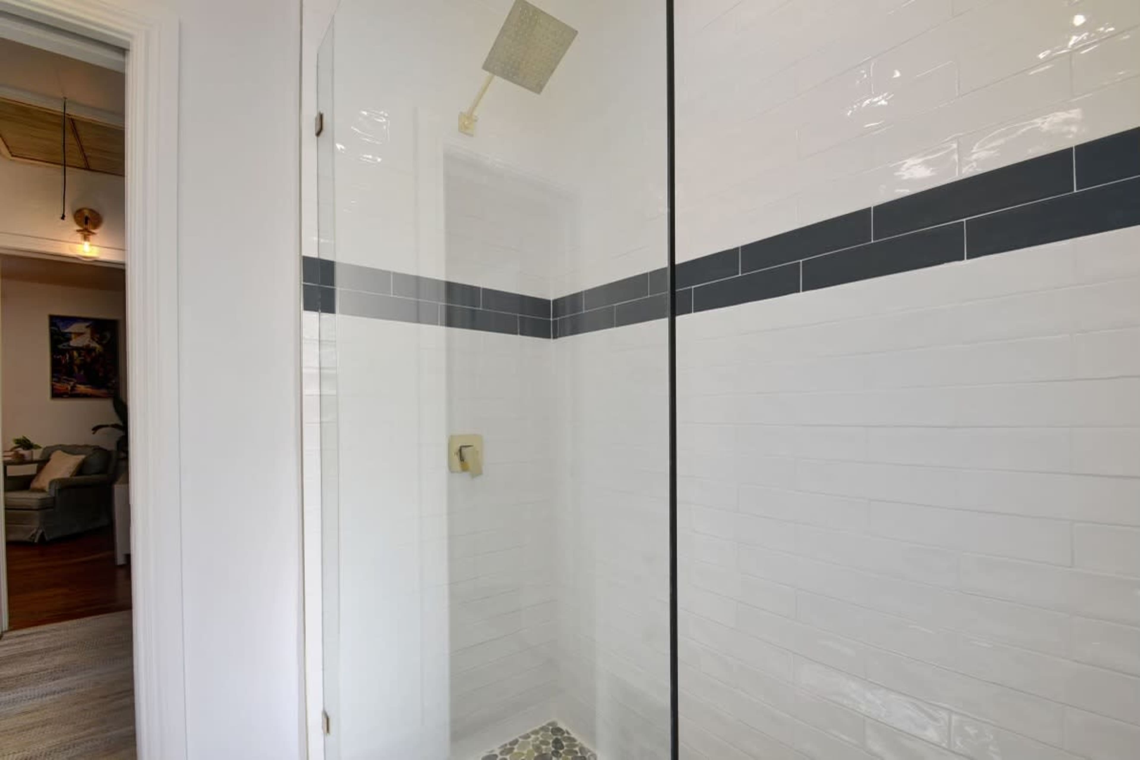 A glass-enclosed shower features white tiled walls with a gray accent band and a pebble tile floor.