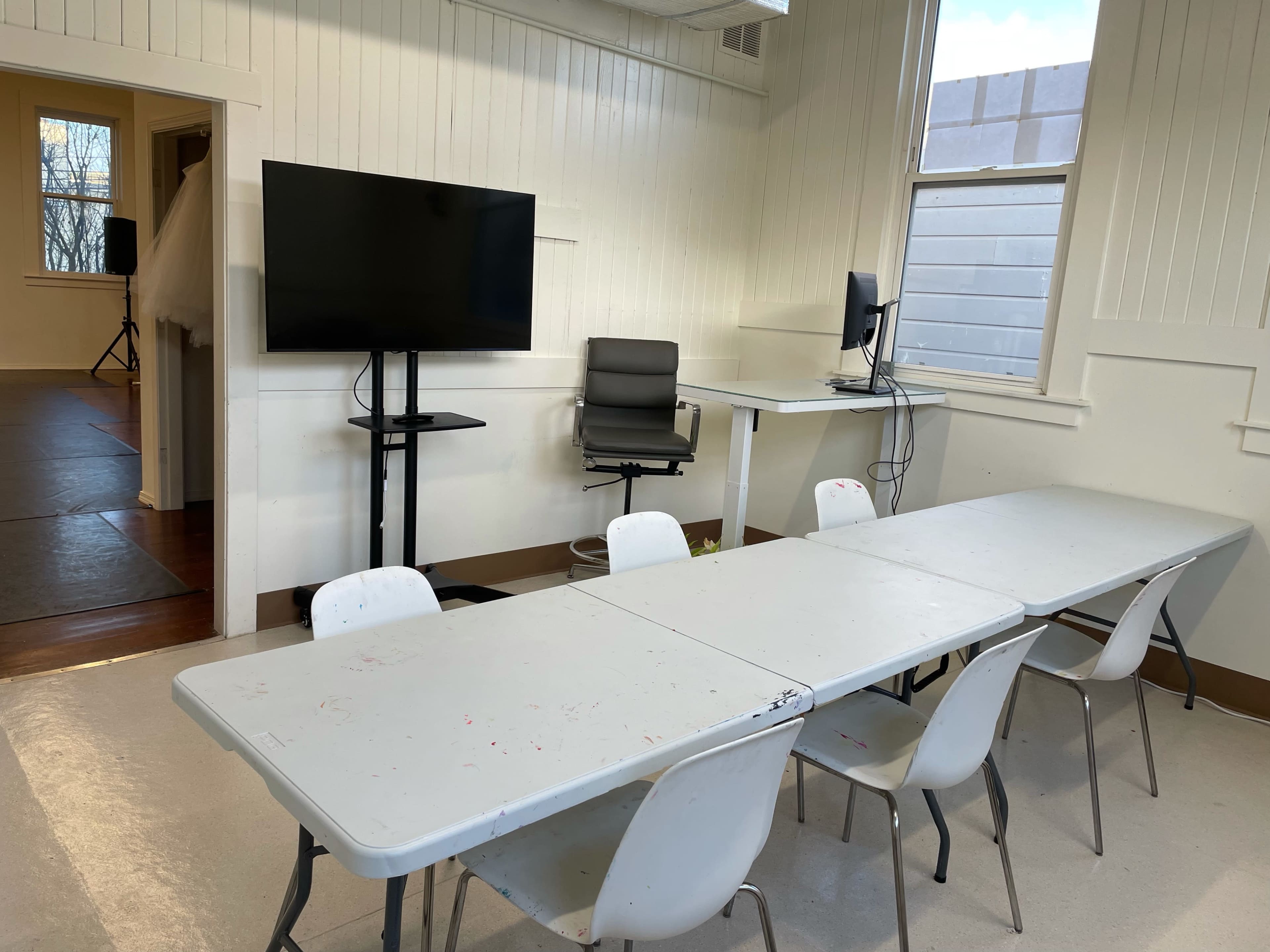 A classroom or meeting room with a long table surrounded by chairs, a television on a stand, and a desk with a computer setup near a window.