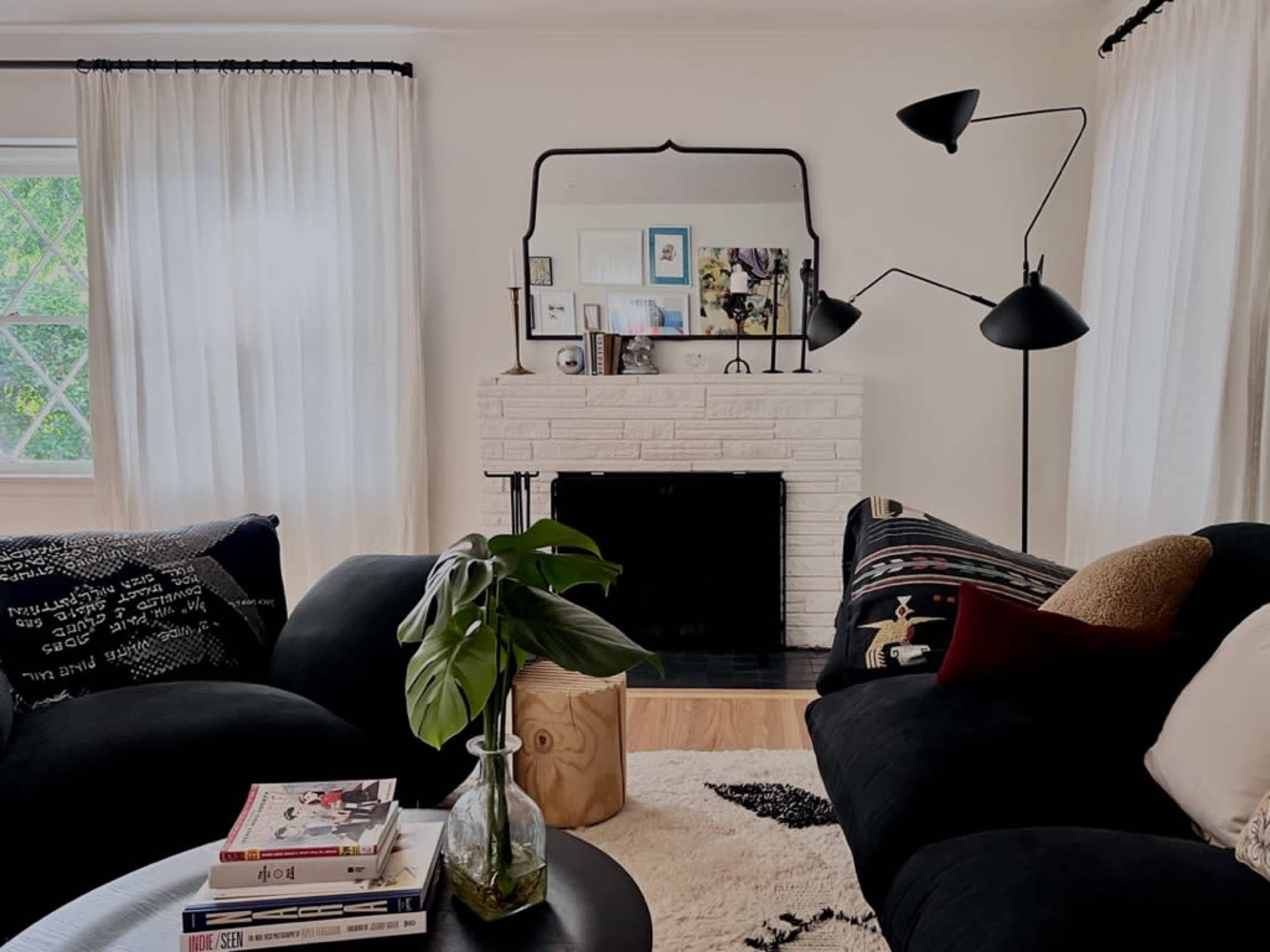 A cozy living room features a white brick fireplace, a large black sofa, and floor-to-ceiling curtains, with a coffee table stacked with books and a potted plant.
