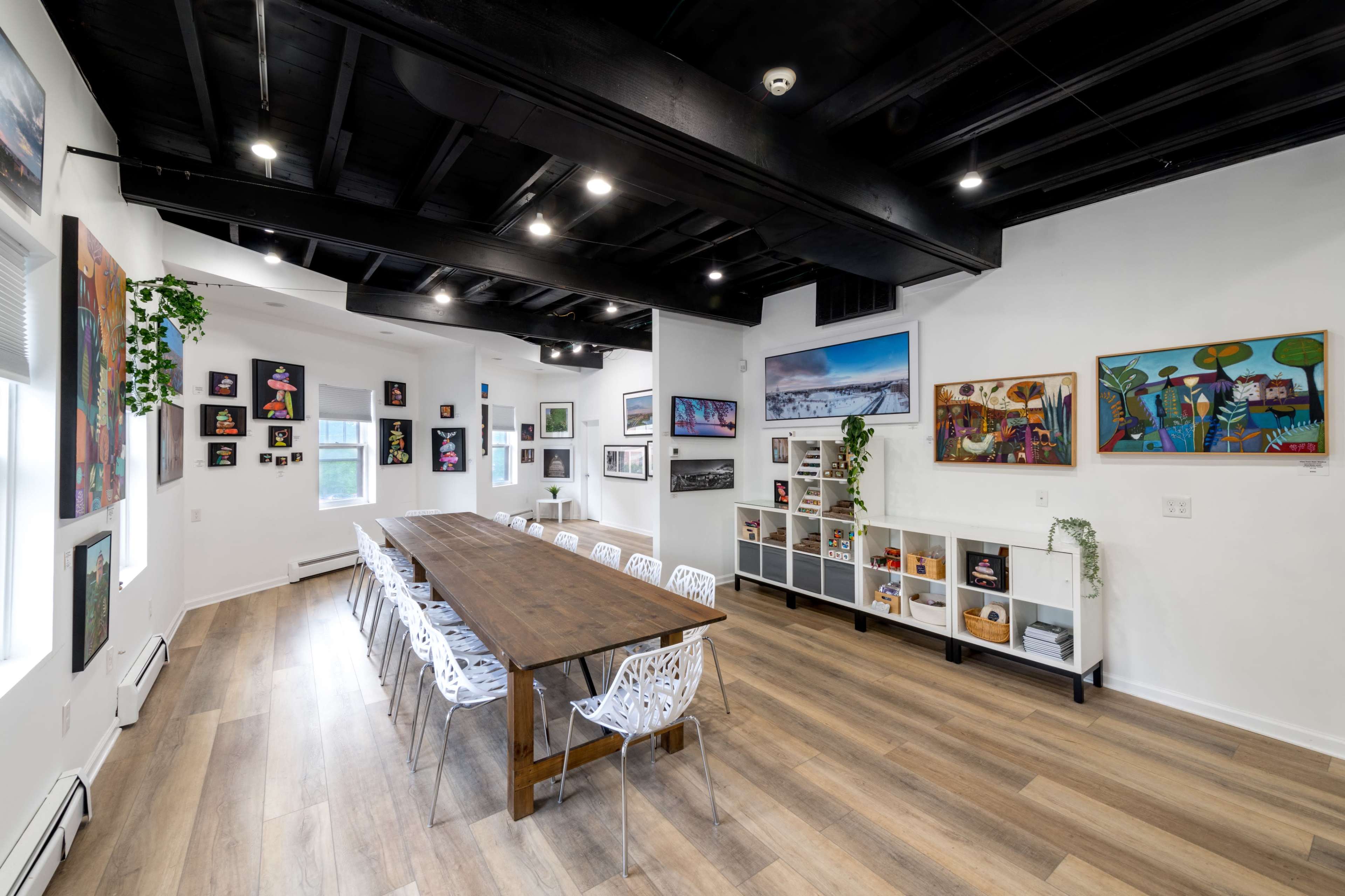 A spacious meeting room with a large wooden table surrounded by white chairs, adorned with artwork on the walls and natural light coming through the windows.