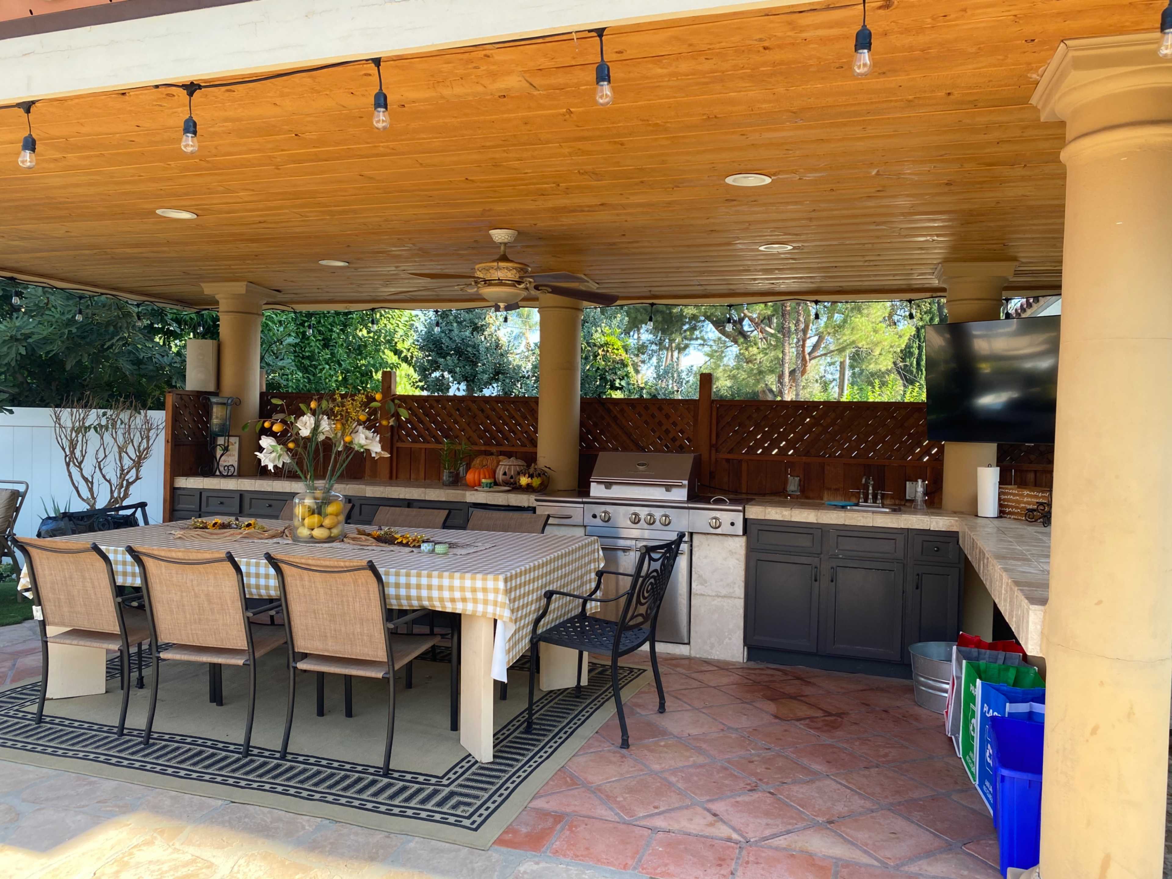 An outdoor kitchen area with a dining table, a grill, and a television, all under a covered roof with wooden beams.