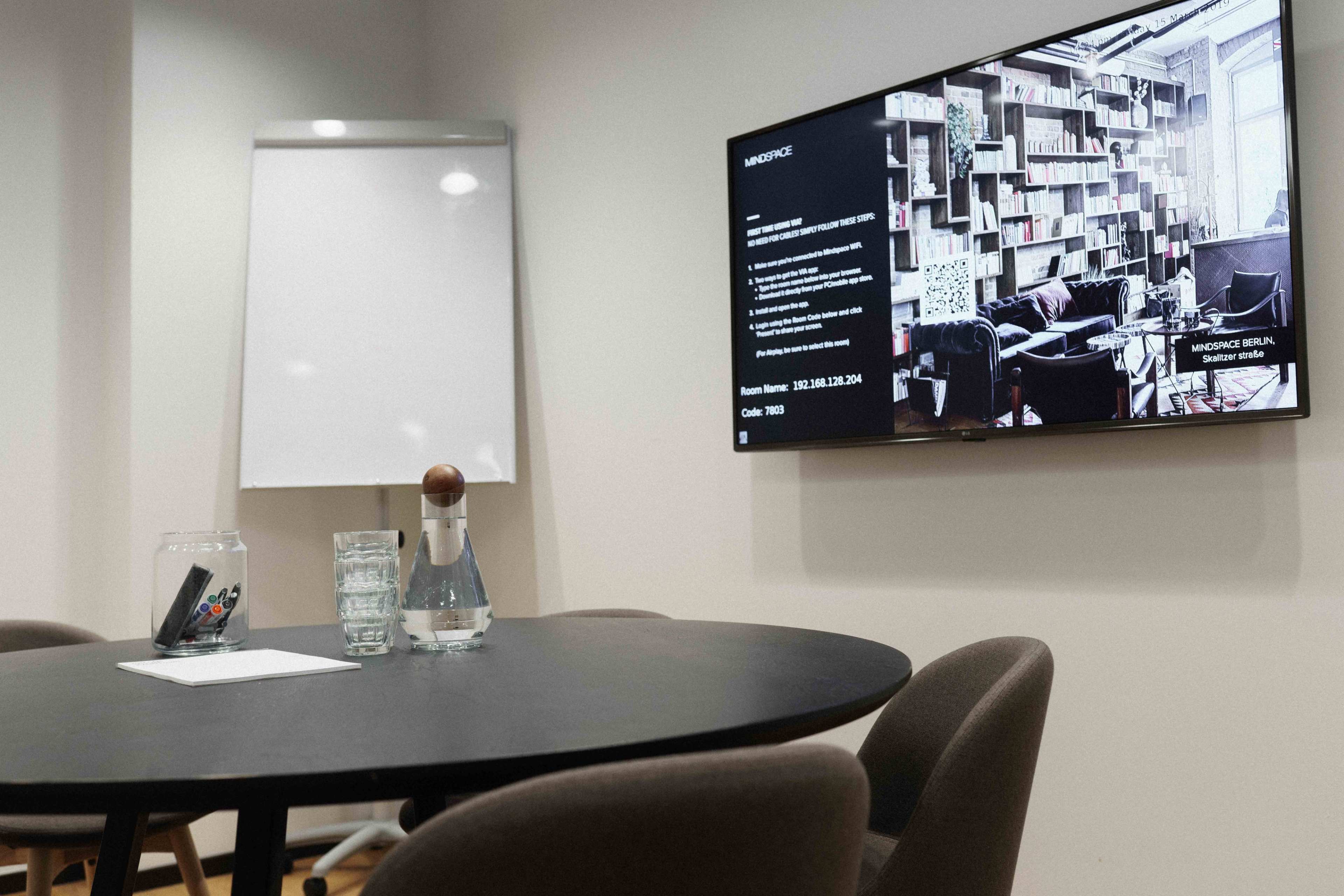 A conference room features a round black table with chairs, a glass water pitcher, and a large screen displaying information, alongside a whiteboard in the background.
