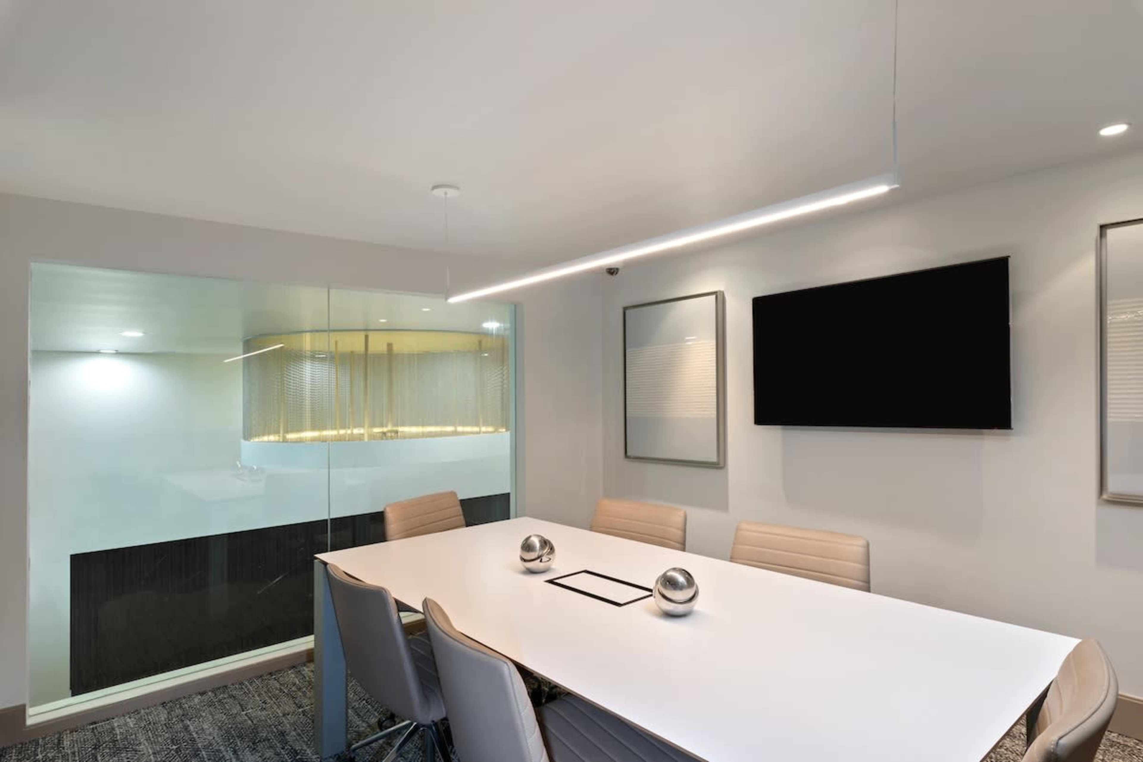 A modern conference room features a large white table surrounded by gray chairs, with a wall-mounted television and a glass partition leading to another space.
