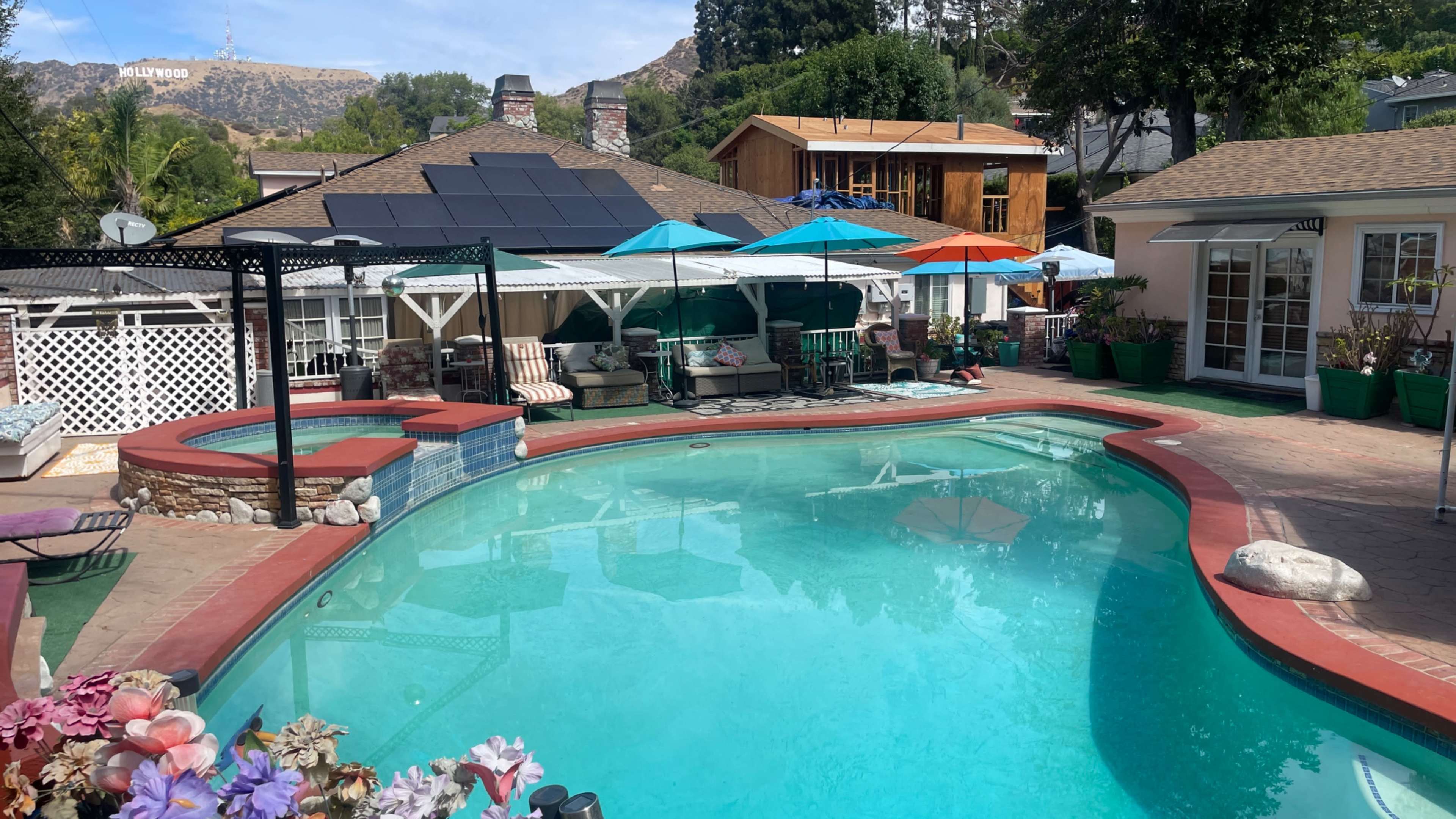 The image shows a residential backyard with a blue swimming pool surrounded by a patio, various seating areas, and several umbrellas, with the Hollywood sign visible in the background.