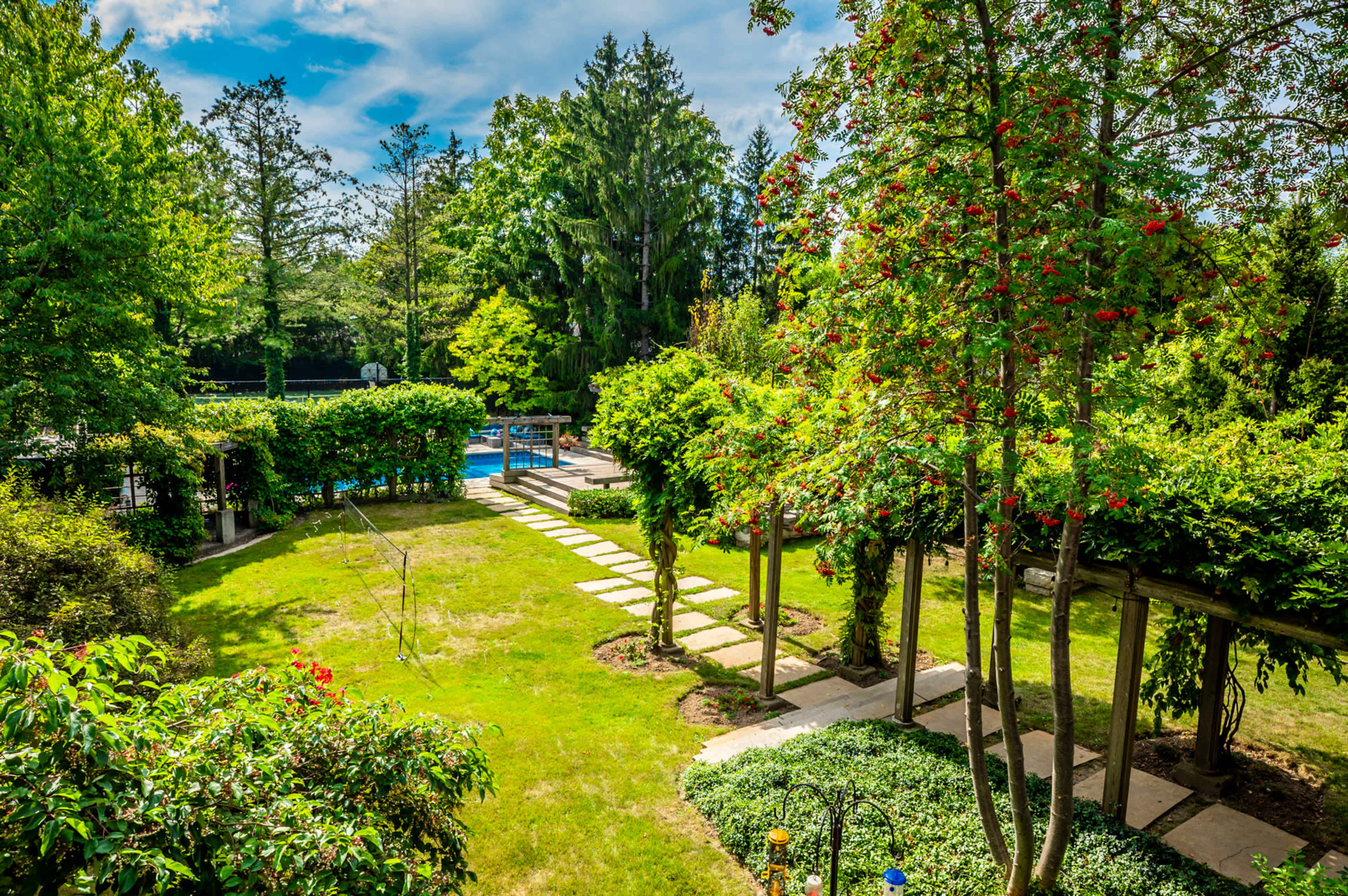 A landscaped garden featuring a stone pathway leading to a swimming pool, surrounded by greenery and trees.