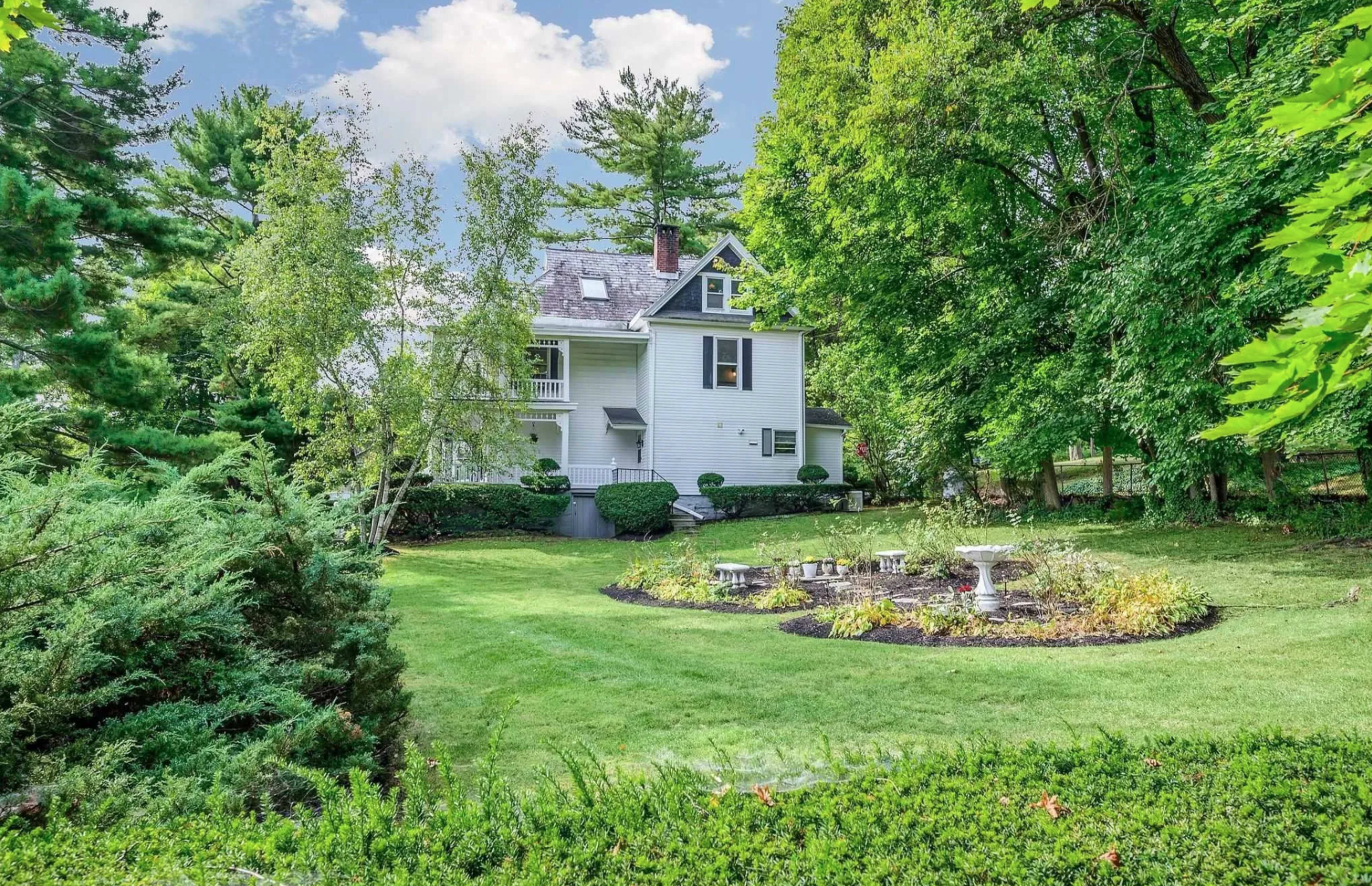 A two-story house is nestled among lush greenery, with a circular garden featuring a stone seating area in the foreground.