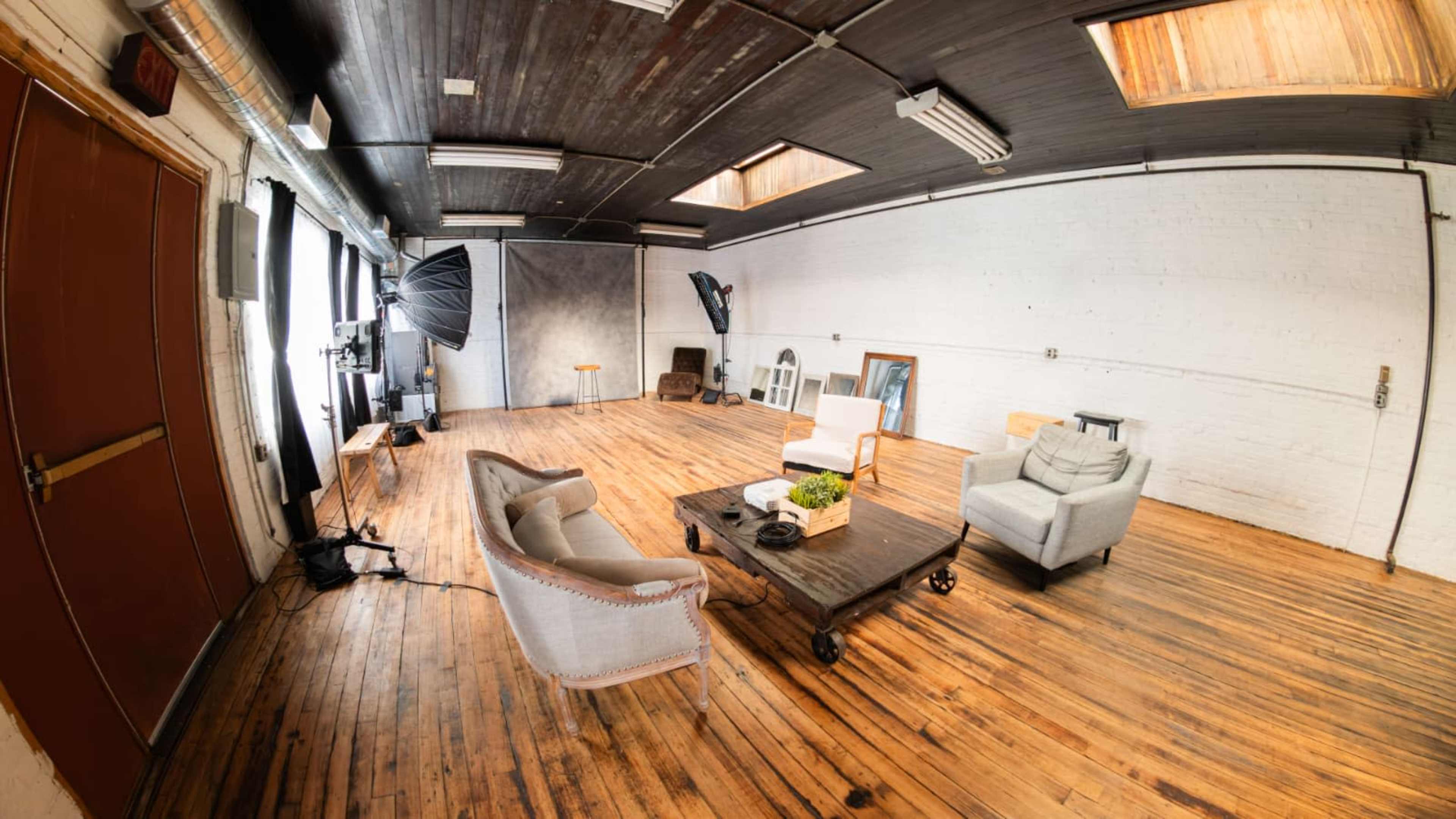 The image shows a spacious studio with wooden floors, two couches, a coffee table, and various photography equipment set up against a plain white wall.