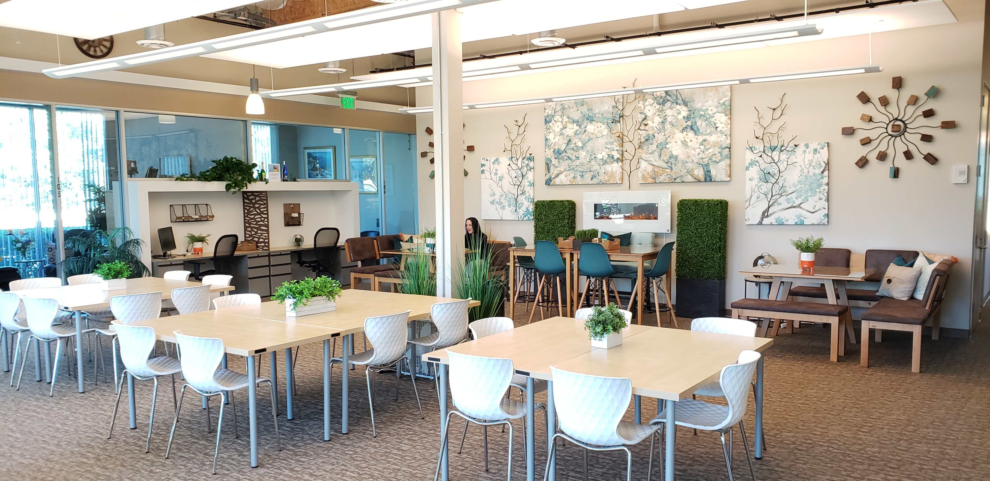 A large, well-lit office space features several tables and chairs, decorative plants, and wall art depicting trees.