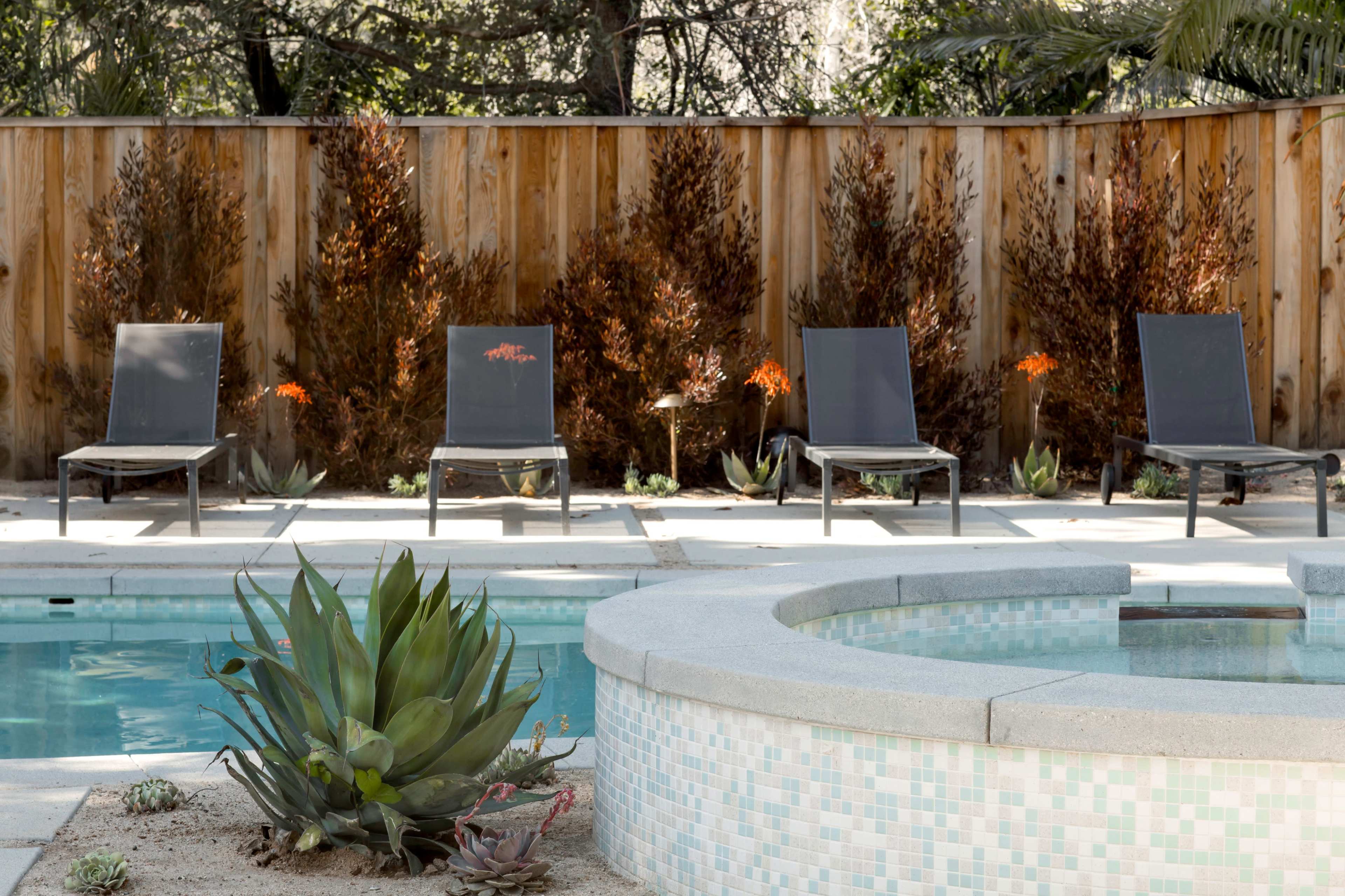 A backyard pool area featuring four lounge chairs, a circular hot tub, and landscaped greenery along a wooden fence.